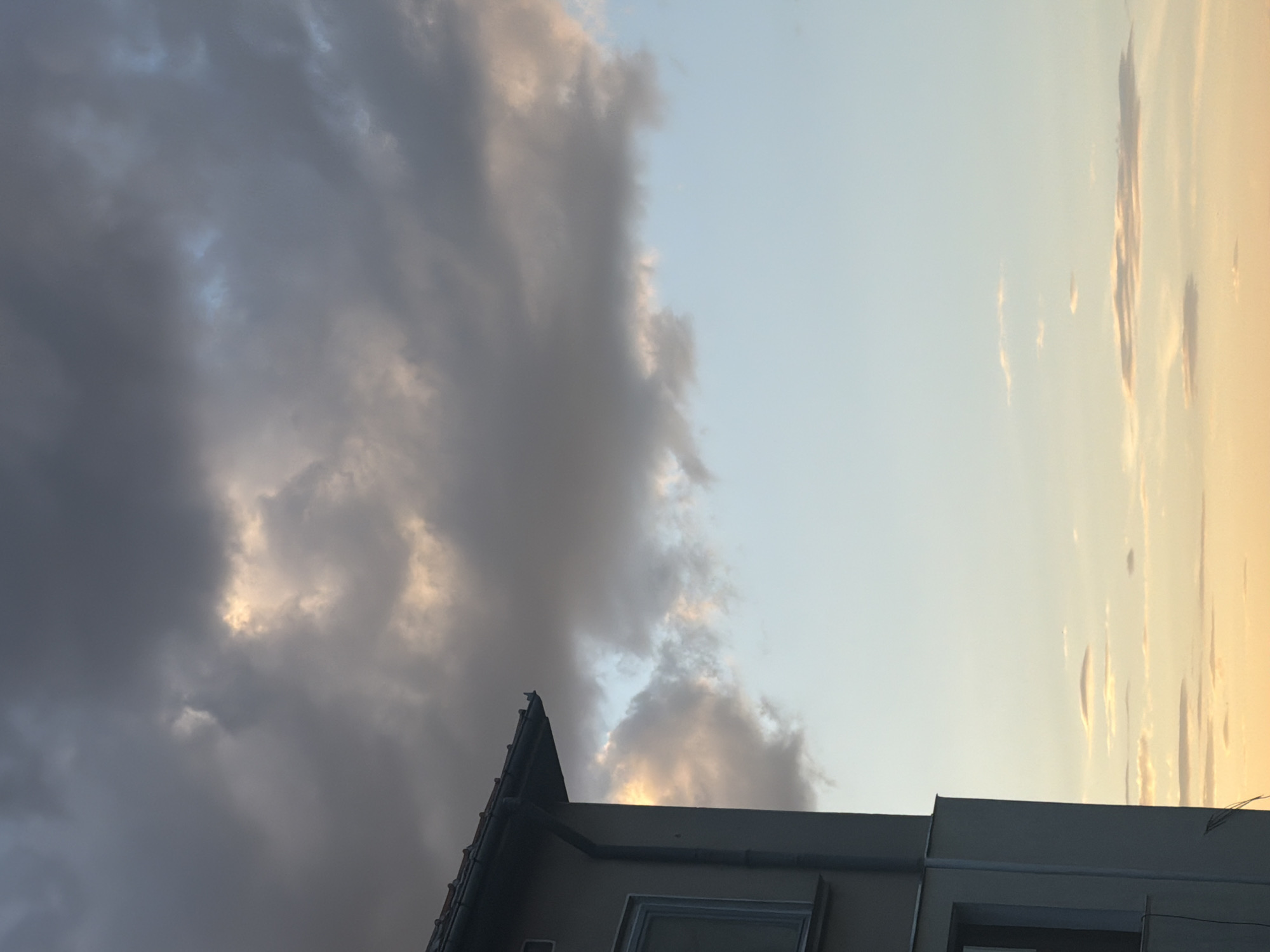 The image shows a building facade on the left, with a grey downspout running vertically along its side. The building is light-colored and has at least one window visible. The roofline extends from the top left of the frame. The majority of the image is occupied by the sky, which displays a mix of clouds. The upper portion of the sky is filled with large, dark grey clouds interspersed with areas of lighter grey and hints of yellow and orange, suggesting the presence of sunlight breaking through. Toward the horizon, the sky transitions to a lighter blue, with scattered, thin, horizontal clouds tinged with yellow and orange, indicative of sunset or sunrise. There are no people or visible actions depicted in the image. The scene is likely set in Belogradchik, Bulgaria, as specified, with the building and sky being the only discernible elements.