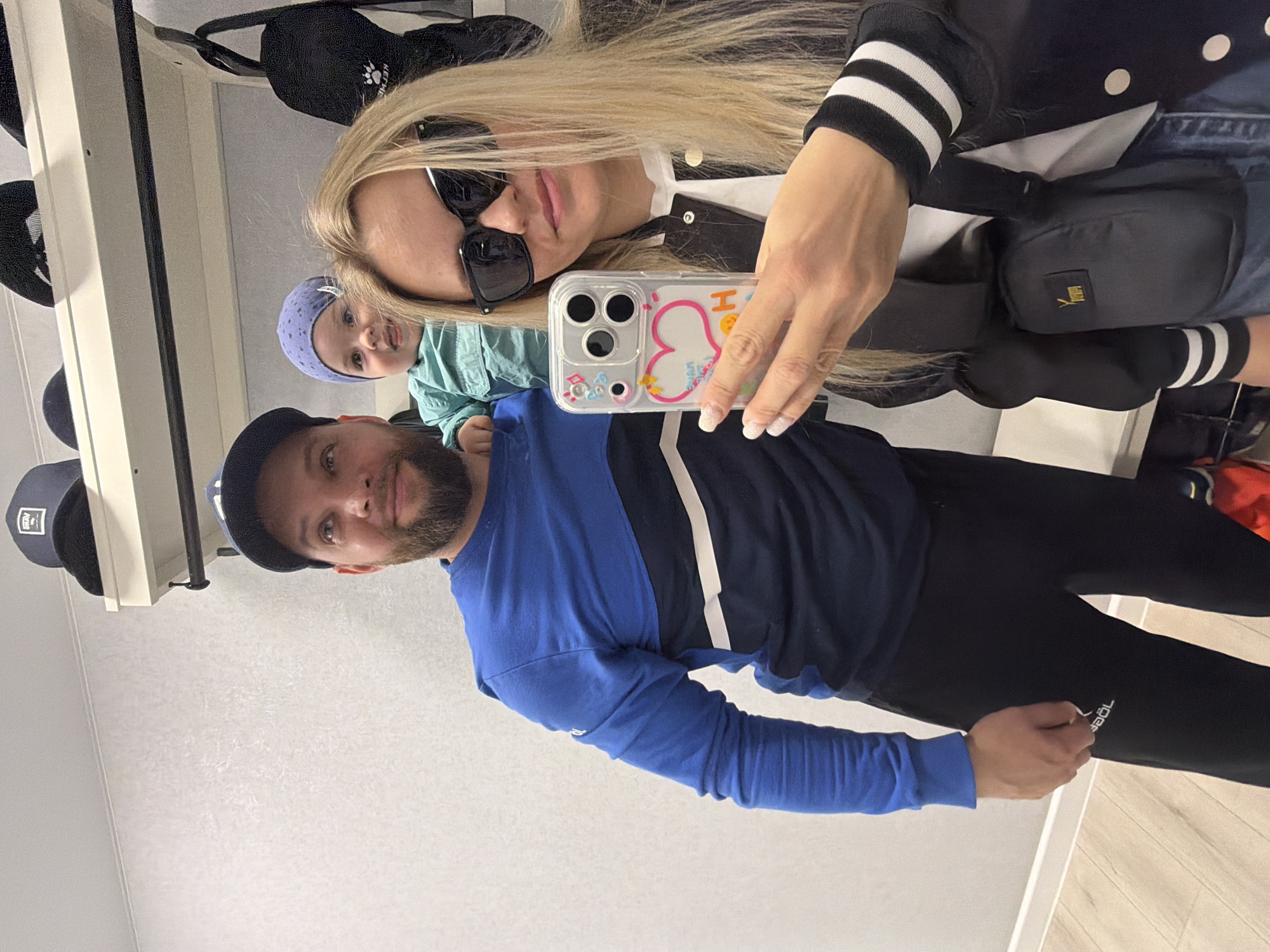 Three people are visible in an indoor photograph. A man with a beard and wearing a blue long-sleeved shirt, black pants, and a black baseball cap appears to be standing next to a woman with long blonde hair and sunglasses who is holding a smartphone to take a selfie. A young child in a blue hat and a light blue garment is visible between the man and woman. The woman is wearing a jacket with white stripes on the sleeves and a black bag across her torso. Hats are hung on a rack in the background. The room has light-colored walls. The location is inferred to be Votkinsk, Russia.