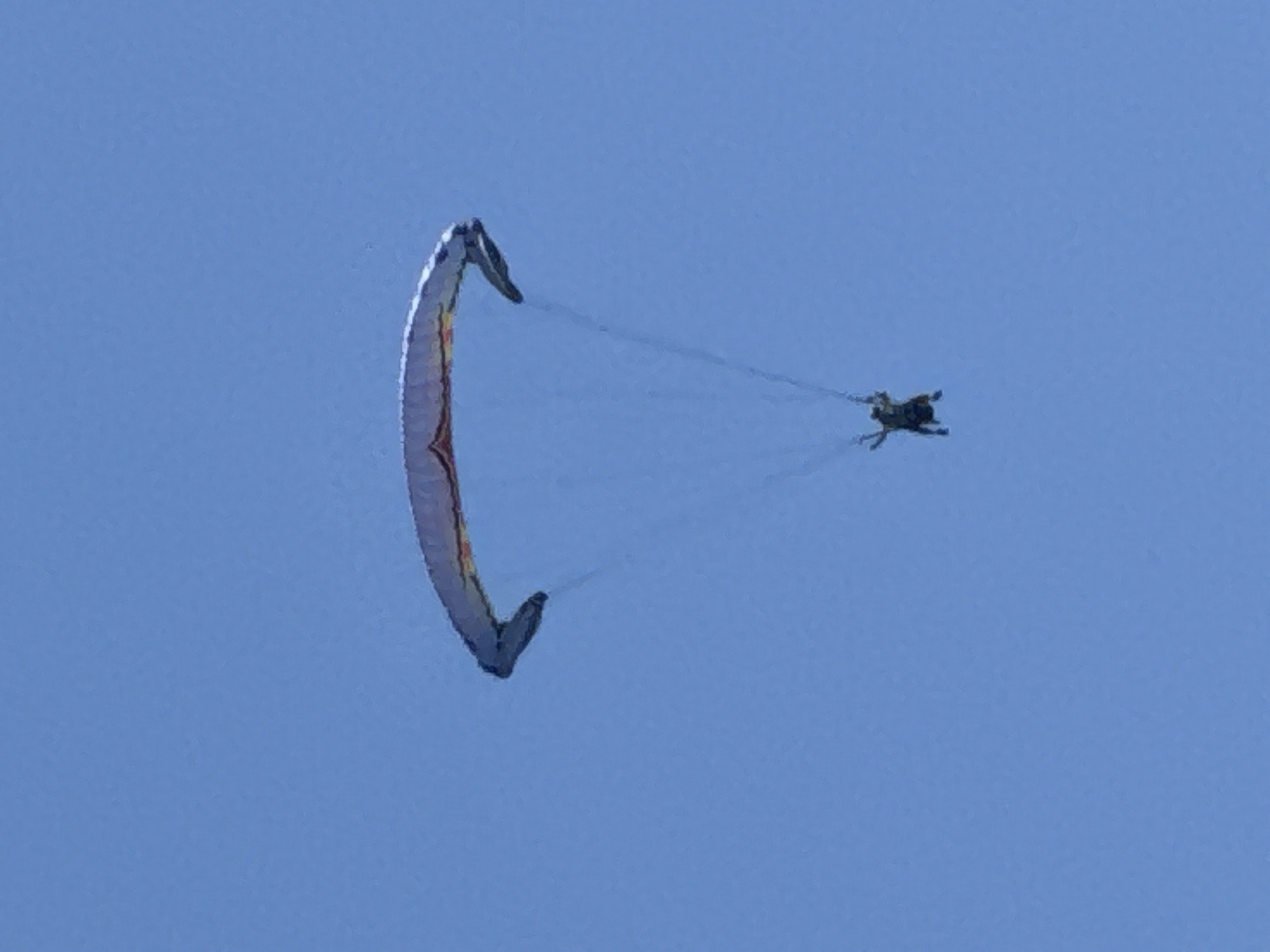 A paraglider with a person suspended below it is captured against a clear blue sky. The paraglider, which appears to be predominantly white with colorful accents, is spread wide and taut. Thin lines connect the paraglider to the person. The individual below is wearing dark clothing and is positioned in a relaxed, upright posture. The scene is devoid of any urban or natural landmarks, suggesting it is an aerial activity taking place in an open, clear sky, with no specific location within Boreti, Montenegro, identifiable from the image.
