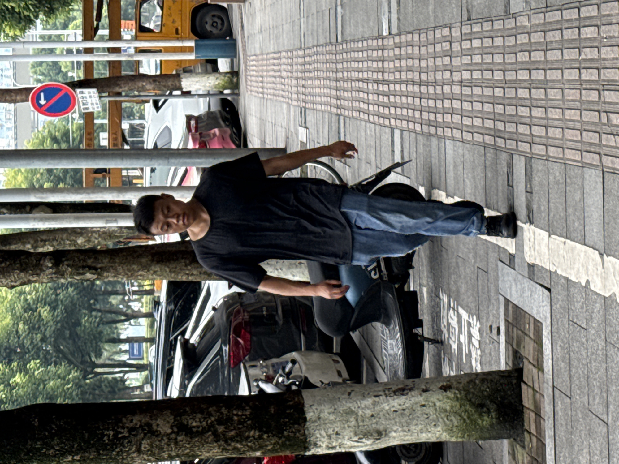 A full shot captures a man walking on a sidewalk in Jiangjin, China. He is wearing a black t-shirt, blue jeans, and black shoes. He is walking towards the camera from the left, with his left hand by his side and his right arm bent at the elbow, possibly holding something. The man is walking on a tactile paving path that runs parallel to a street lined with trees and parked cars. A blue scooter is parked to the left of the man, and a yellow truck is visible in the background to the right. A traffic sign is also visible on a pole to the right of the man. The day appears overcast.