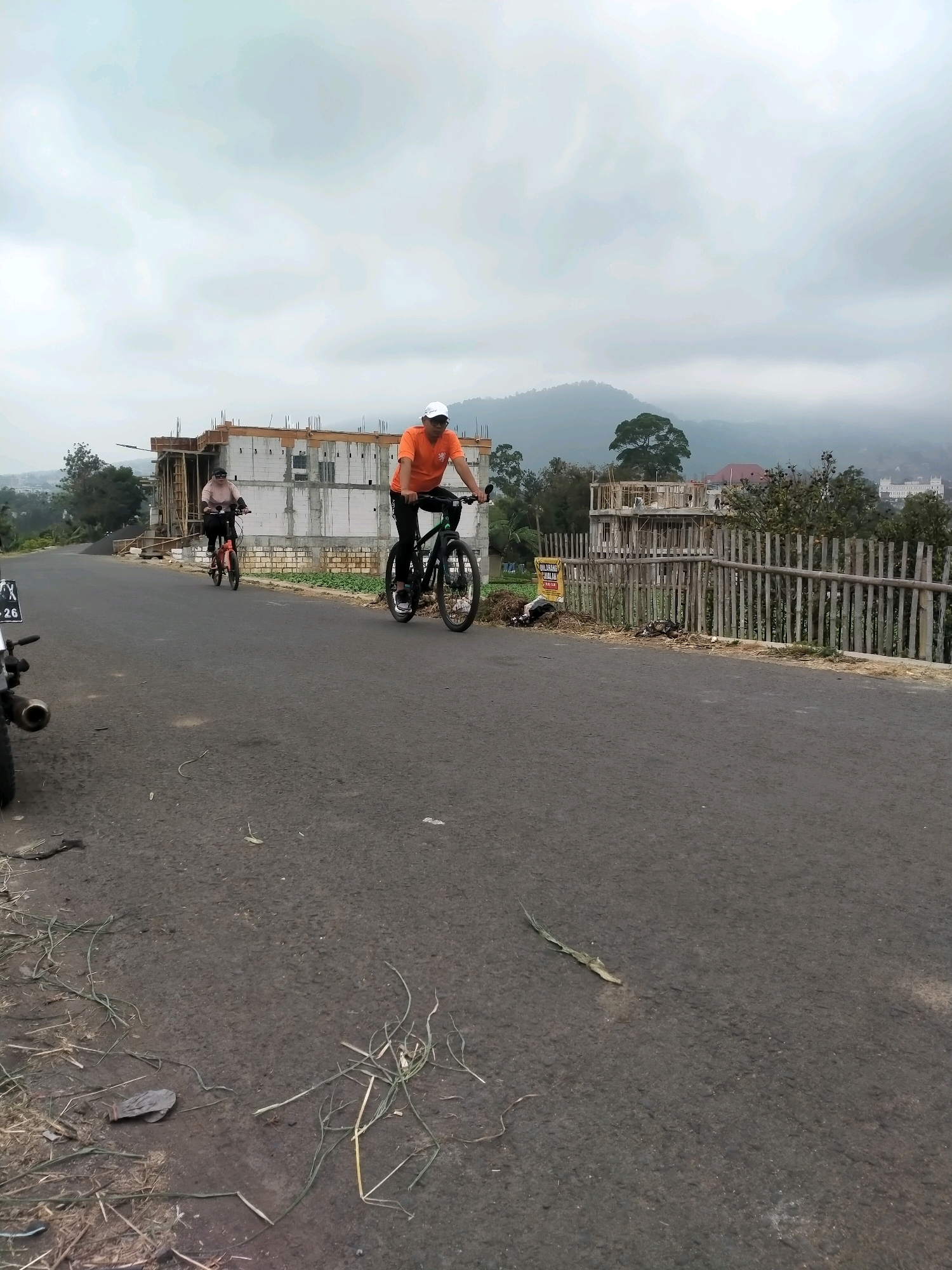 In Batu, Indonesia, on an overcast day, two individuals are cycling on an asphalt road. The person in the foreground, wearing an orange t-shirt and a white cap, is on a black bicycle. Behind them, another person in a light pink shirt and hijab is riding a red bicycle. To the left, a motorcycle is partially visible. In the background, a multi-story building under construction is seen, with scaffolding and brickwork. A wooden fence runs along the side of the road, and beyond it, lush green trees and a distant, mist-shrouded mountain range are visible. The road has scattered debris, including grass and a discarded glove.