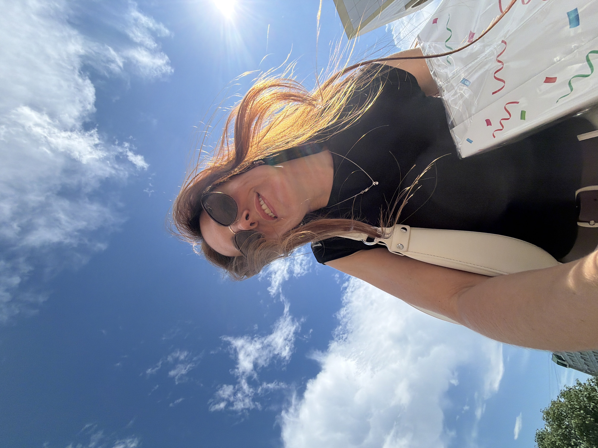 A woman with long, reddish-brown hair and sunglasses is taking a selfie outdoors. She is smiling, and her hair is blowing in the wind. She is wearing a black top and a light-colored purse is slung over her shoulder. In the background, the sky is a vibrant blue with scattered white clouds. The sun is shining brightly, creating a lens flare in the upper left corner of the image. There's a hint of a building and some green trees in the lower right corner. The overall impression is one of a bright, sunny day. The media file appears to be a casual, candid shot.