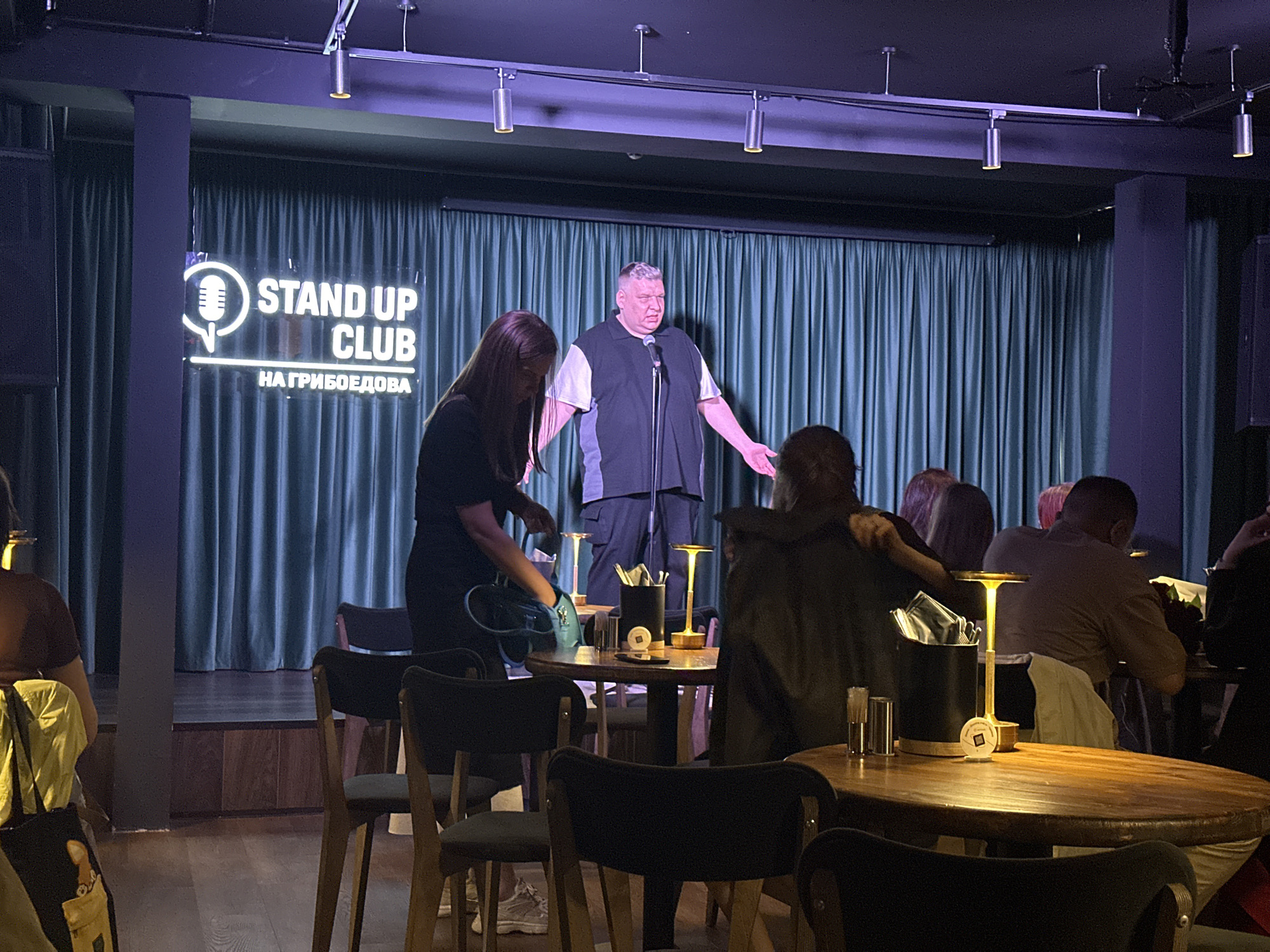The image shows a stand-up comedy performance taking place at the "Stand Up Club Griboedova" in Saint Petersburg, Russia. A man, likely the comedian, stands center stage with a microphone. In the foreground, audience members are seated at tables, some engaged in actions like a woman accessing a purse. The stage is set against a backdrop of blue curtains, with the club's neon sign visible on the left side. Cylindrical lights are hanging from the ceiling. The lighting is dim with spotlights on the stage.
