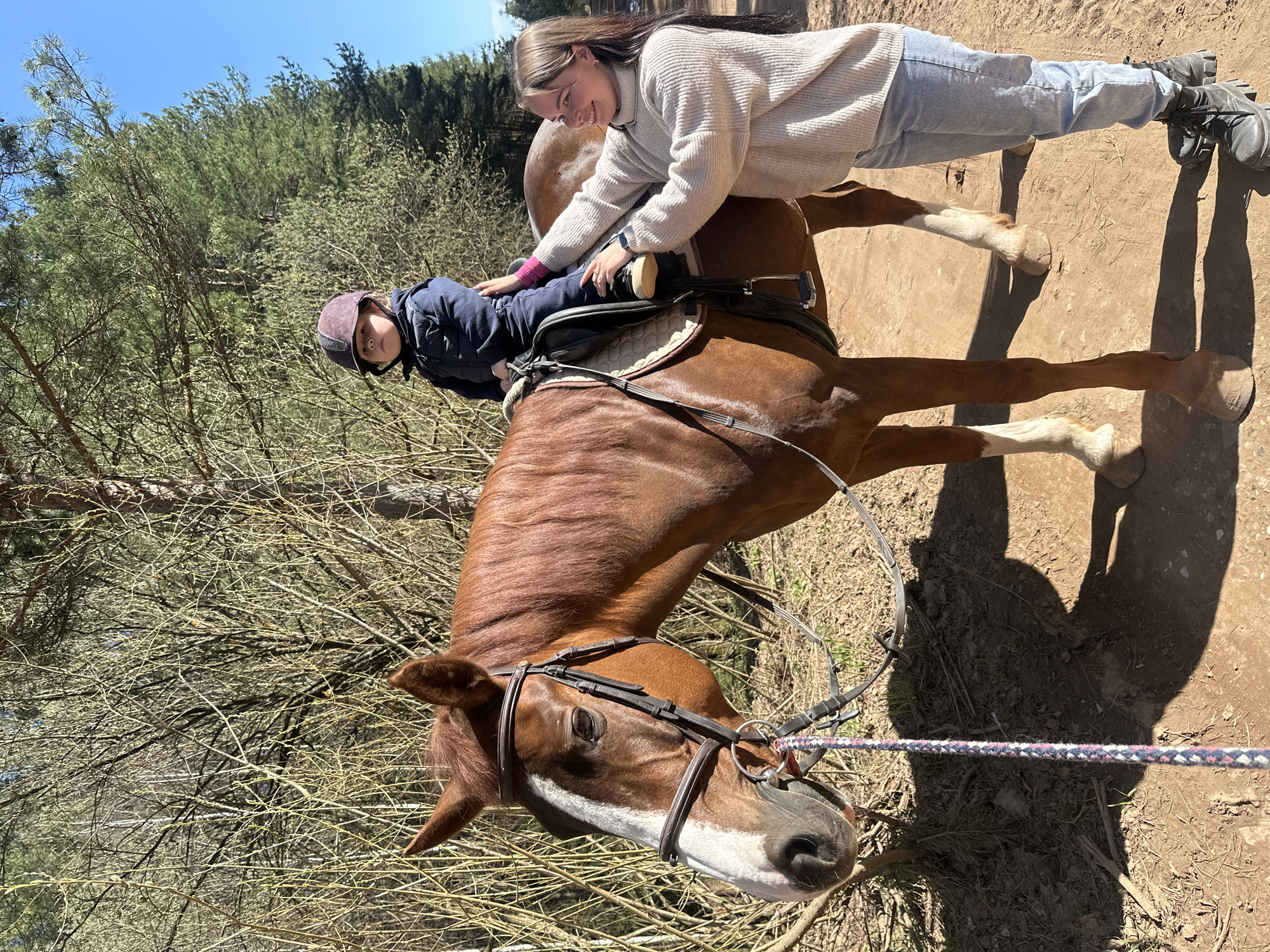 A medium shot depicts a brown horse with a white blaze standing in a dirt area near trees. A child wearing a riding helmet and blue jacket is sitting in a saddle on the horse. A young woman in a light-colored sweater and jeans is standing next to the horse, holding onto the child. The ground is dry with dirt, and there are shadows visible. The background consists of trees and a clear blue sky. The location is inferred from the query to be Русский Вожой, Russia.