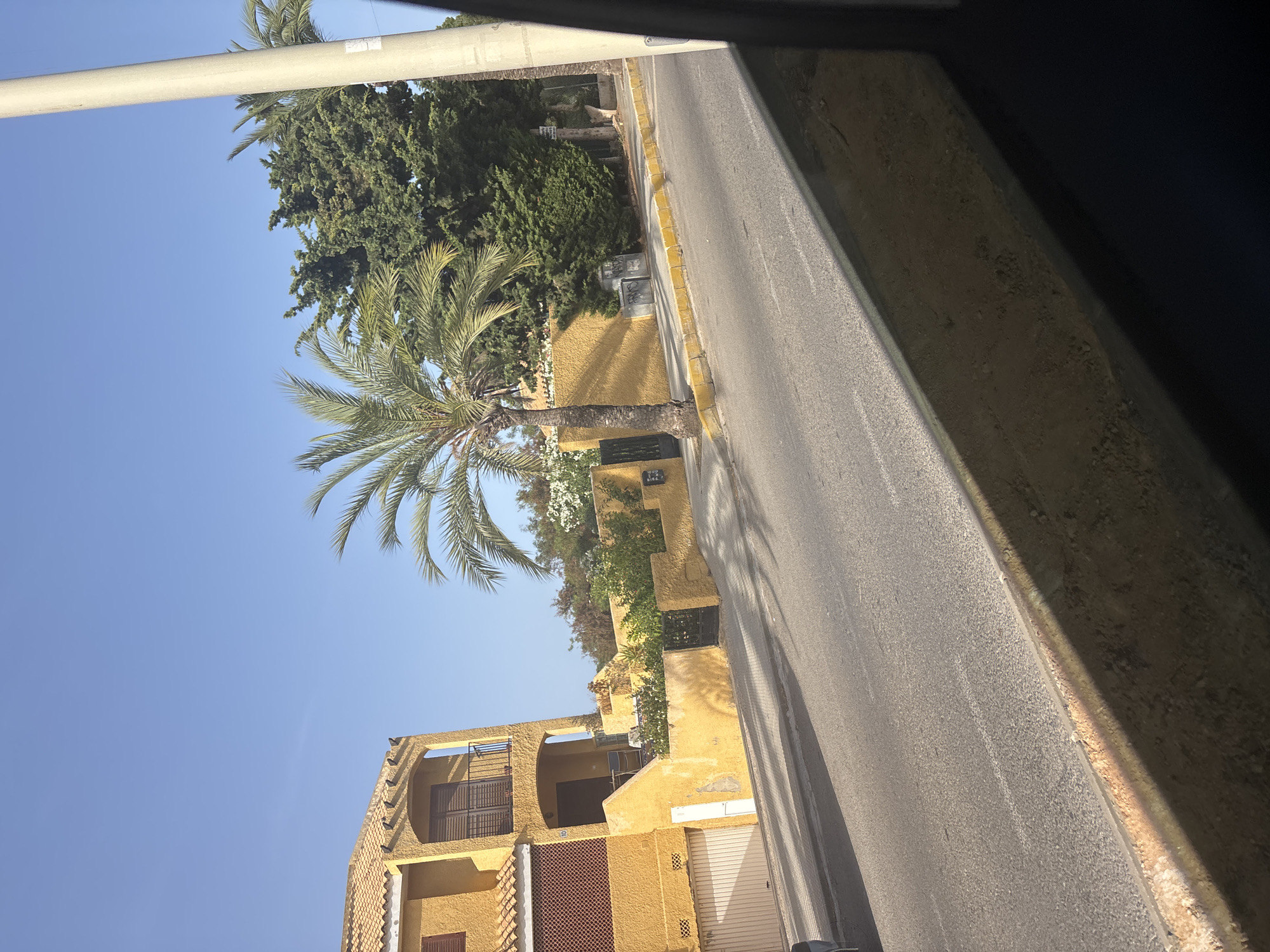 The image shows a yellow house with a tiled roof on the left, a palm tree in the center, and a street in the foreground. The house has a balcony with dark railings and brown shutters on the windows. There are garages on the ground floor. A yellow wall surrounds the property, with a black metal gate. Green trees are visible behind the house. The street is paved and has a yellow curb. The sky is clear and blue. A vertical light pole is on the right side of the image. The image is taken from a moving vehicle, as indicated by the blur and the angle.