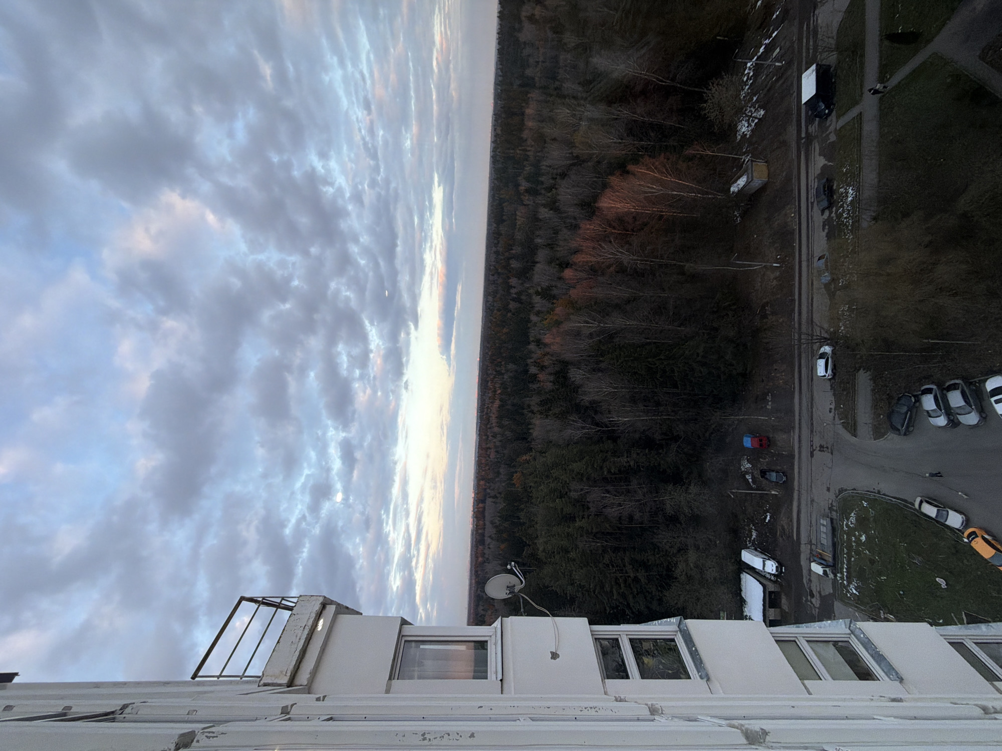 The image presents an expansive, high-angle view, likely captured from a multi-story building, overlooking a mixed landscape during either sunrise or sunset. The original image is vertically oriented, but the content depicts a horizontal vista.
The upper portion of the scene is dominated by a cloudy sky displaying varying shades of grey and blue, with streaks of soft pink and orange hues along the distant horizon, indicative of either dawn or dusk. A faint crescent moon is visible in the upper-mid section of the sky.
A broad band of dark, dense forest stretches across the mid-ground, acting as a natural divider. The trees appear mostly bare or with minimal foliage, suggesting late autumn, winter, or early spring.
Below the forest, the scene transitions to human infrastructure. Roads, asphalted parking areas, and sparse grassy patches are visible. Numerous parked cars of various colors, including white, red, black, and a distinctive yellow/orange vehicle, are scattered along the roadsides and in lots. Other objects include streetlights and what appears to be utility structures or containers.
In the immediate foreground, elements of the viewpoint building are visible: a white railing, parts of white window frames, and a white satellite dish attached to the building structure.
No specific activity or event is taking place; the scene is a static portrayal of a landscape at a particular time of day. No individuals are clearly discernible. The weather appears overcast but calm. There is no visible text in the image. The vast forest, combined with the residential/urban structures, is consistent with a location such as Brekhovo, Russia, experiencing cooler seasons.