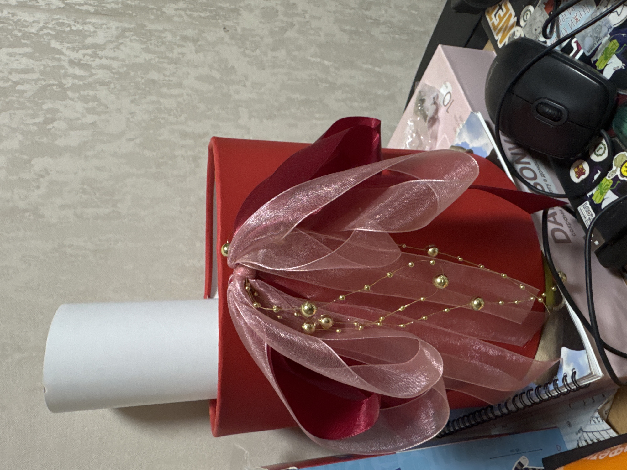 The image displays a red gift box or packaging, oriented horizontally, on a light-colored, textured surface. A prominent decorative bow is attached to the top of the red box. This bow is fashioned from two types of ribbon: a solid, dark red satin ribbon and a sheer, shimmery pink or light red organza ribbon. Woven into the bow and draped across the top of the red box is a string of small golden beads or pearls.
Protruding from the left side of the red box is a white, cylindrical object, possibly a rolled-up paper, document, or a tube. The background areas, particularly to the right and upper right, reveal a cluttered desk or table setting. Visible items include a black computer mouse with a scroll wheel, a pink or light purple box, and a spiral-bound notebook. Various stickers are adhered to the surface behind these objects. Partially visible text on the stickers and background includes fragments of what appears to be Cyrillic script (e.g., "ПЕРВЫЙ К", "ДА") and English words such as "SONY", "ON USE", and "NOVE". The lighting suggests an indoor setting, and no specific time of day or weather is discernible. No people are present, and no active event is taking place; the scene is a static arrangement of objects.