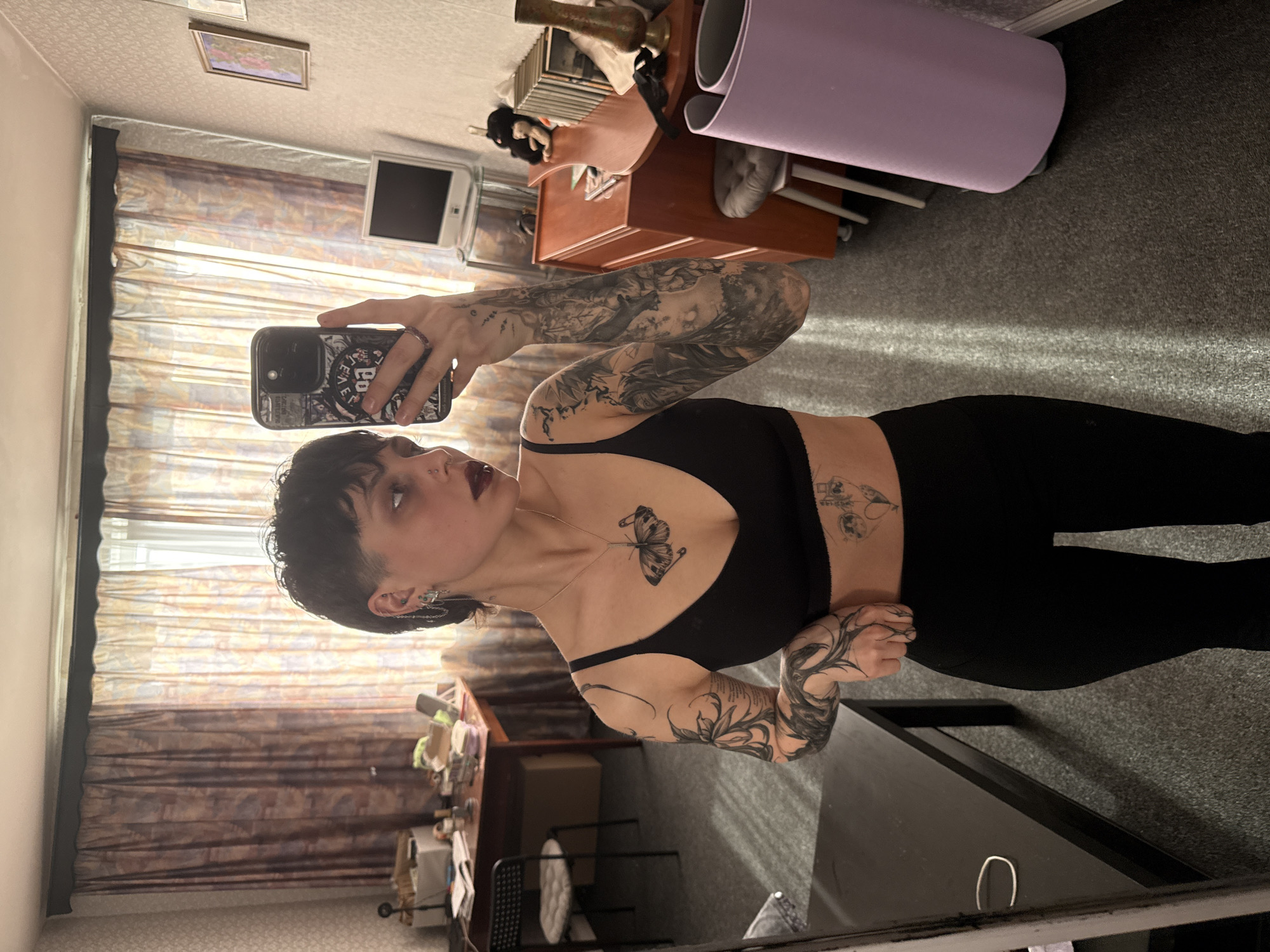 A slender woman with dark hair and multiple tattoos is taking a selfie indoors. She is wearing a black sports bra and black leggings. Her phone case is black with a design on the back. A butterfly tattoo is visible on her chest.
The room has a window with patterned curtains. A wooden desk with various items on it, including a white monitor, is present. A rolled-up purple yoga mat rests against the wall. The floor is carpeted in gray.
Due to the limited information and angle of the shot, precise identification of the location in Saint Petersburg is not possible.