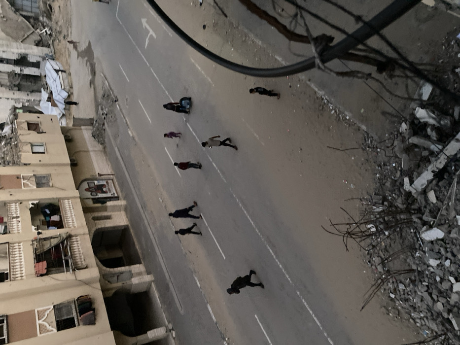This image depicts an urban street scene in Khan Yunis, Palestinian Territory, viewed from an elevated perspective. The scene is characterized by active human presence amidst significant structural damage.
In the foreground, a large pile of concrete rubble, fragmented blocks, and twisted rebar is visible, indicating a destroyed structure. A thick, dark cable runs vertically on the far right, partially obscuring the view.
The midground features a paved two-lane road, showing areas of dust and sand. White solid lines mark the lanes, and a white arrow indicating forward movement is painted on the right lane.
Several individuals are present on the street:
- Two adults in dark clothing walk side-by-side from left to right on the left lane.
- Another adult, wearing a red top and dark pants, pushes a dark, wheeled object across the street from left to right.
- An adult in a light-colored top and dark pants walks from right to left, appearing to look at a mobile phone.
- A small child in a pink outfit stands near the center of the road.
- Another small child in dark clothing stands on the right side of the road, closer to the foreground.
- In the background of the street, a figure in dark clothing walks away from the viewer.
To the left of the street, a multi-story building, light brown or beige in color, stands largely intact. It features balconies with white balustrades, some with laundry hanging. Arched entrances are visible on the ground floor, where a red and white sign is also mounted.
To the right of the street and extending into the background, widespread destruction is evident. Buildings are heavily damaged or partially collapsed, presenting exposed internal structures, piles of debris, and twisted metal. A large piece of white material, possibly a tarp or corrugated metal, lies amidst the wreckage.
The lighting is soft and even, suggesting an overcast day or the twilight hours.