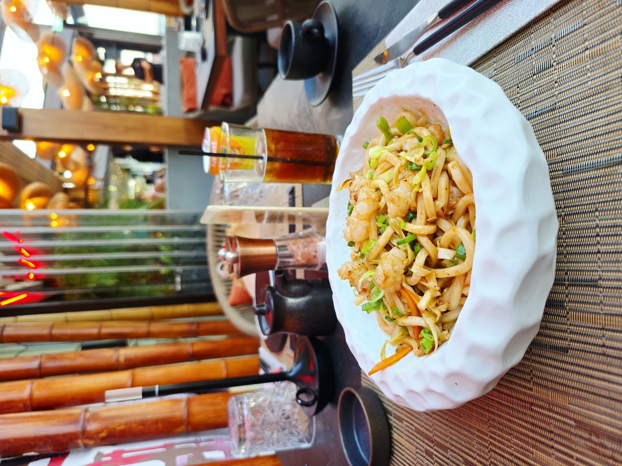 A close-up, eye-level shot captures a dish of noodles with shrimp and vegetables, presented in a white, intricately textured bowl. The bowl sits on a woven placemat, accompanied by silverware and a small dipping dish. In the background, a glass of iced tea and a can of soda are visible, along with a black cup and saucer. The scene is set in a restaurant with a bamboo-themed decor, featuring tall bamboo stalks and warm, ambient lighting. The overall atmosphere suggests a casual dining experience.
