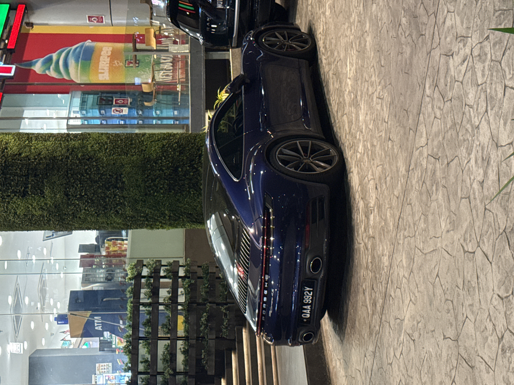 The image shows a dark blue Porsche 911 Carrera 4S parked on a paved area. The vehicle's rear is prominently visible, displaying the license plate "QAA 992Y." The backdrop features a 7-Eleven store, recognizable by its Slurpee advertisement and storefront. The scene is located in Kuala Lumpur, Malaysia. A hedge of greenery separates the store from the parked vehicle. Several wooden structures can also be seen near the ATM store.