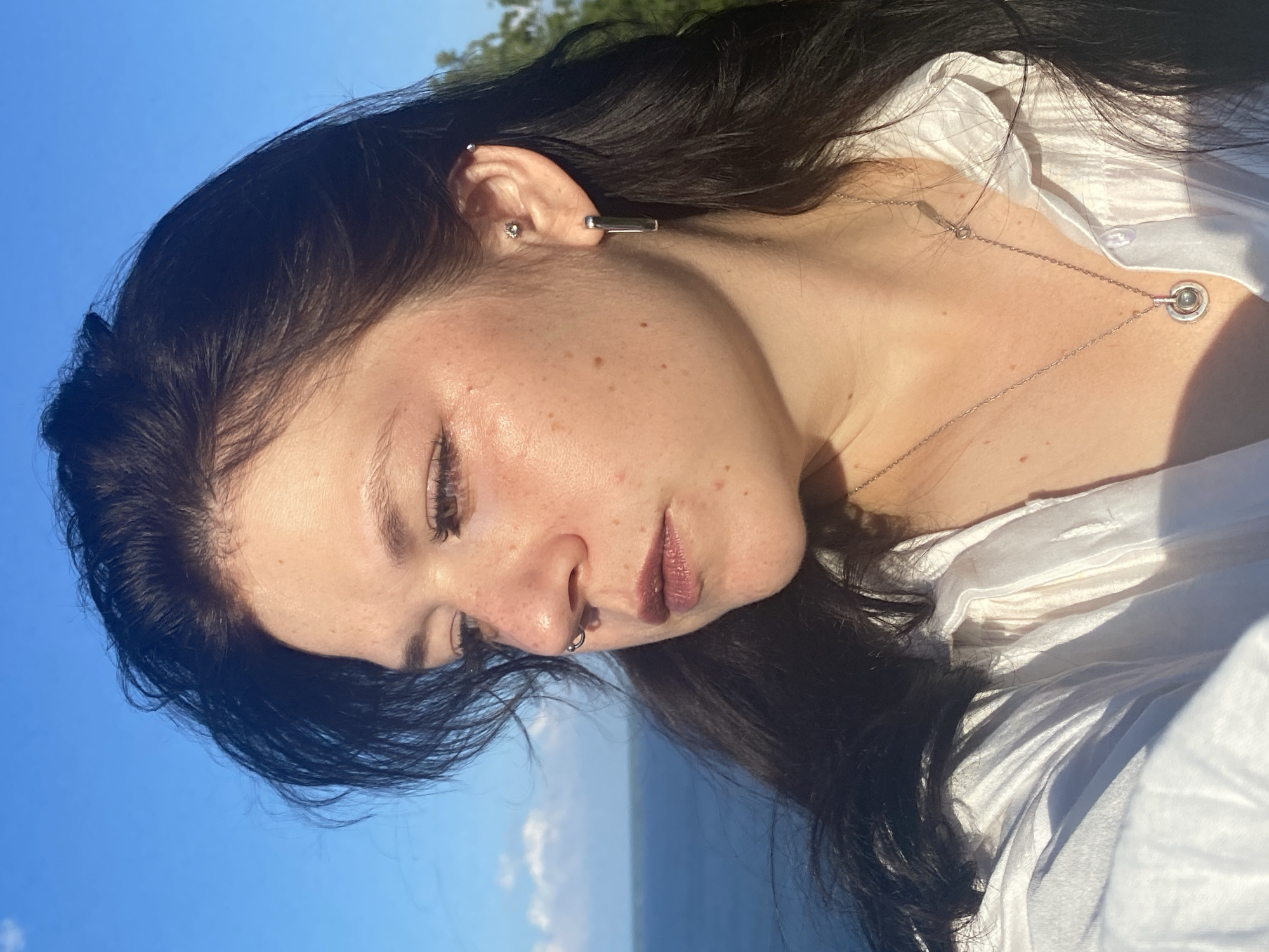 A medium shot captures a young woman with dark hair, freckles, and subtle makeup looking away from the camera. She wears a white collared shirt and a silver necklace with a circular pendant. Her left ear is adorned with two earrings, one small and stud-like, and the other a silver bar. She also has a septum piercing. The background features a hazy blue sky with scattered clouds, a body of water, and the faint outline of trees on the right. The lighting suggests it is daytime, with sunlight casting shadows across her face and shirt. The location is specified as Kostovaty, Russia.