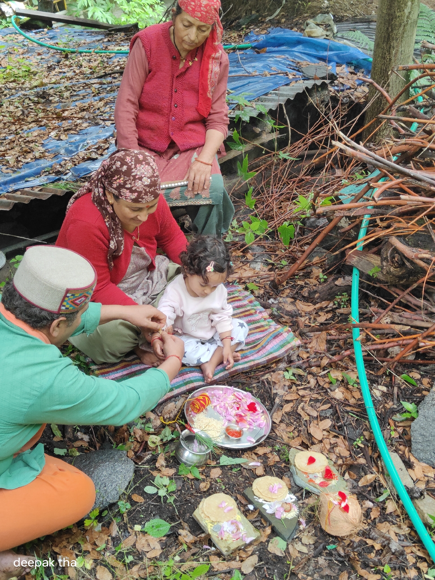 In Vashisht, India, an outdoor scene unfolds with four individuals, two adults and one child, engaged in a ritual. A man, wearing a green shirt and orange pants, with a traditional Himachali cap, is tying a red thread around the wrist of a young child. The child, dressed in a light pink top and white pants, sits on a colorful mat. Beside them, an adult woman, adorned in a red headscarf and a red sweater over a patterned dress, observes the ceremony. Another adult woman, also wearing a patterned headscarf and a red sweater, is seated, looking down. The ground is covered with dry leaves and twigs, suggesting a natural or rural setting. Scattered around are ritualistic items, including a silver plate with rice, flower petals, and a small container of red paste, as well as coconuts and flatbreads on stone slabs. A blue tarp and corrugated metal are visible in the background, along with a pile of branches and a green hose, adding to the rustic environment.