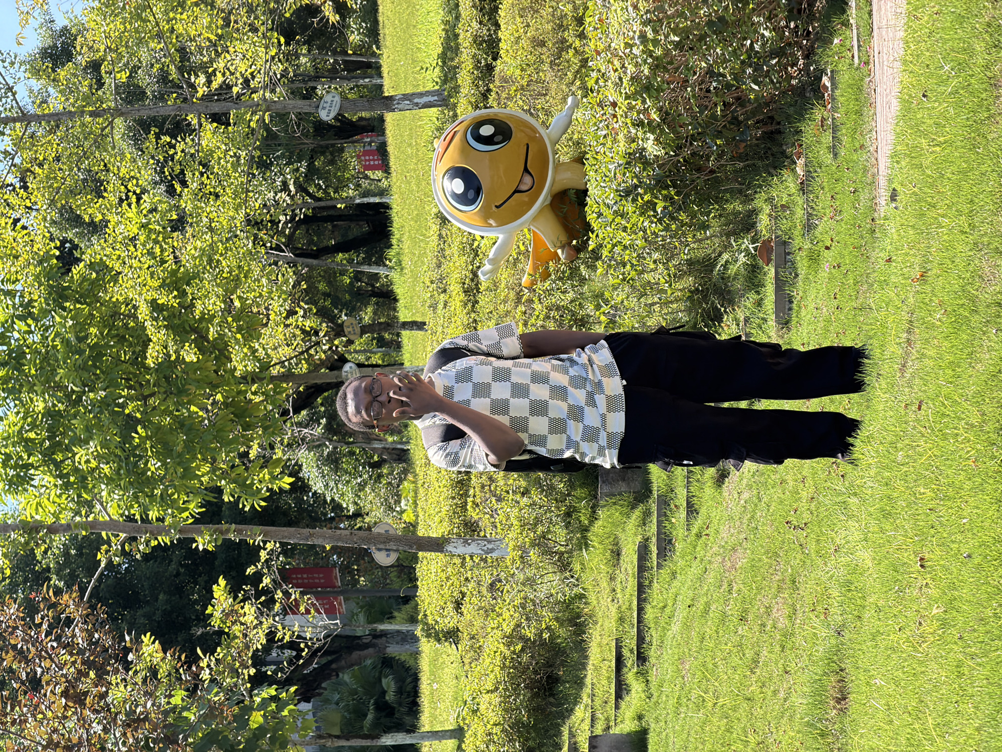 In the city of Jiangjin, China, a young Black male stands on a grassy lawn, holding his hand to his mouth. He is wearing glasses, a checkered t-shirt, and black cargo pants, with a backpack on his shoulders. To his right, a large, cartoonish yellow statue with wide eyes and a smiling mouth is partially obscured by bushes. The background features lush green trees and a bright, sunny sky. Stone steps are visible on the left side of the image, leading up a grassy incline.
