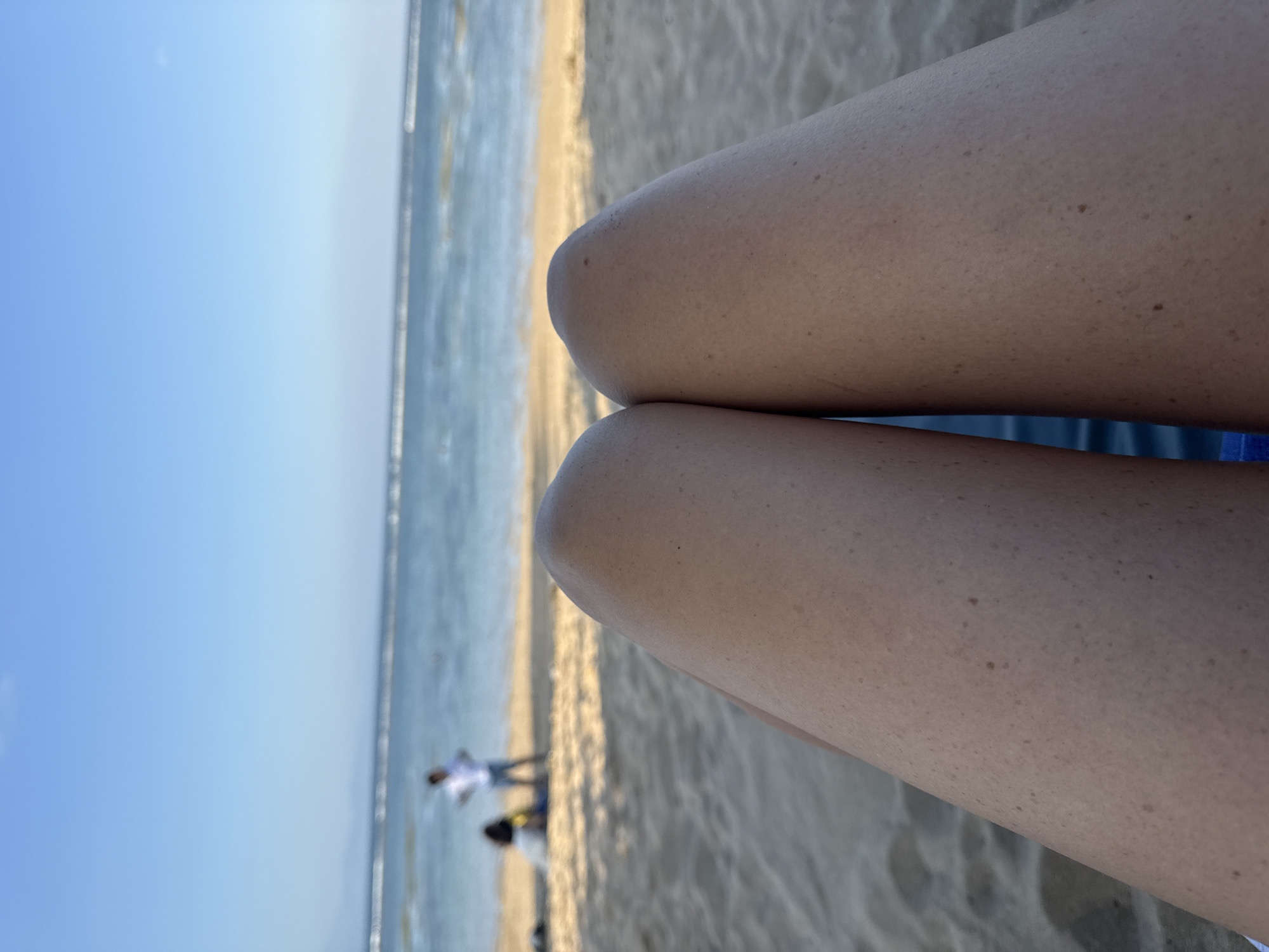 A first-person perspective shot shows a person's legs resting on a beach chair with the ocean and sky in the background. Two people are visible in the distance, one standing and one sitting. The sand is light brown, and the ocean is a calm blue with light waves. The sky is a clear, bright blue. The image appears to have been taken at Kuta Beach in Denpasar, Indonesia.