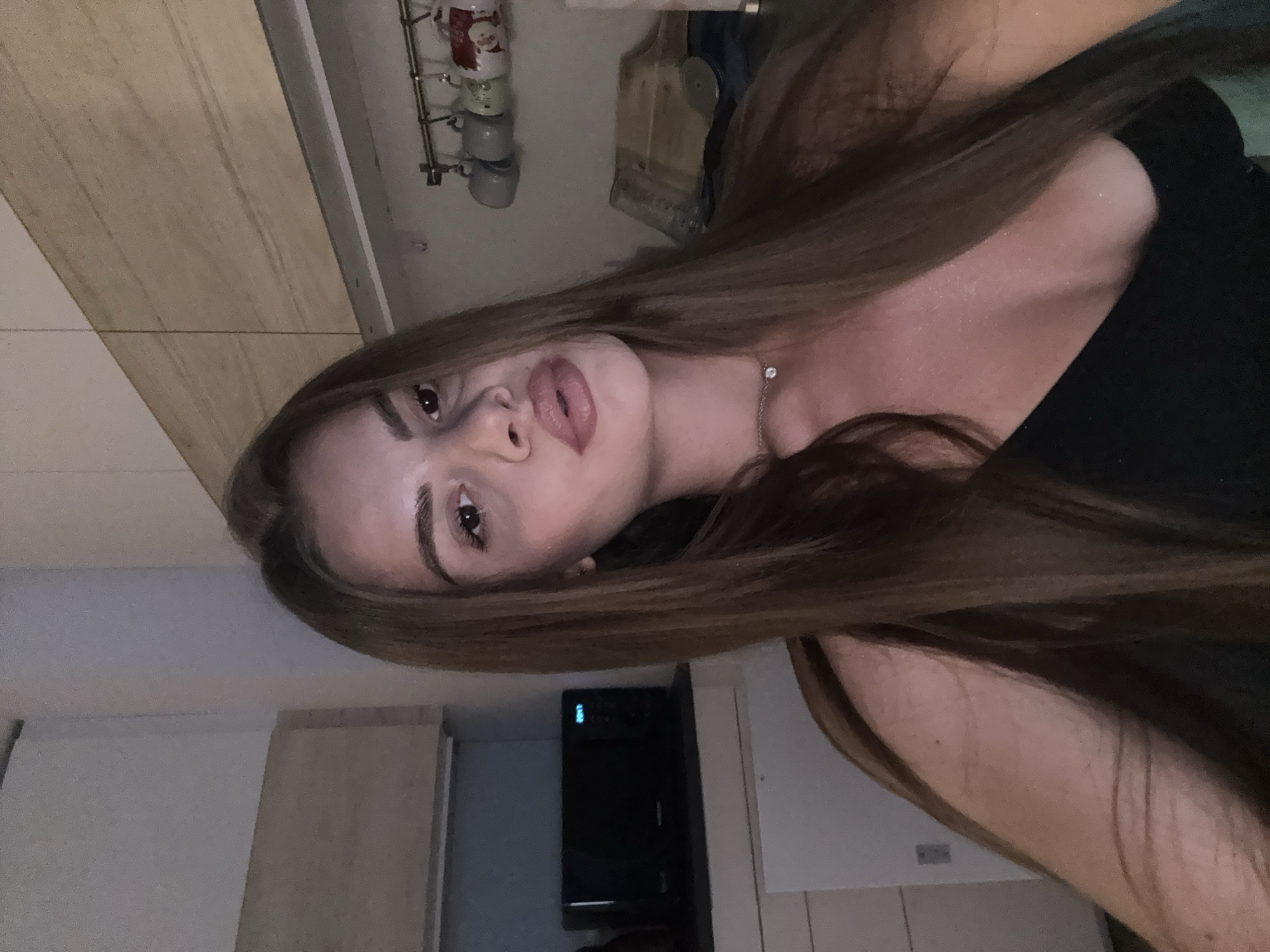 The image features a young woman prominently in a close-up, selfie-style shot, taken indoors, likely in a kitchen or dining area in Plovdiv, Bulgaria. She has long, straight, light brown hair that frames her face and extends over her shoulders. Her dark eyes are directed towards the camera, and her full lips are slightly parted in a pout, accentuated by what appears to be lip gloss or lipstick. She wears a dark, possibly black, top and a delicate silver-colored chain necklace with a small, sparkling pendant at her throat. A subtle shimmer or highlight is visible on her skin, particularly on her décolletage.
The background suggests a domestic setting. To her left, a light-colored wooden surface, possibly a cabinet or wall panel, is visible. Above her head, a metal rack holds several mugs, some decorated with festive patterns, including what looks like Santa Claus. Beside the mugs, a wooden cutting board and a pizza cutter are hanging. Further in the background, to her right, a dark kitchen appliance, possibly a microwave or oven, displays a blue digital "0:00" on its clock. The lighting in the scene is artificial and somewhat dim, consistent with an indoor evening or night setting. The image itself is oriented sideways, with the woman's head tilted to the left.