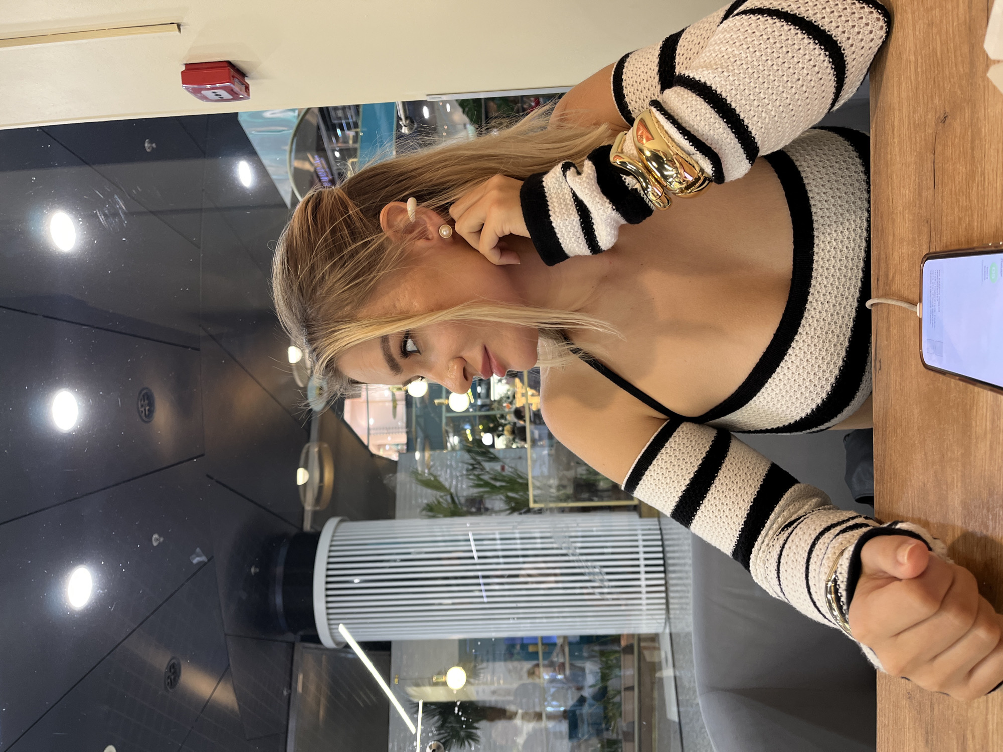A young woman with blonde hair tied up is sitting at a wooden table, looking to her right. She is wearing a black and white striped top with detached sleeves and large gold bracelets on her left wrist. A pearl earring and an ear cuff adorn her right ear. In front of her is a smartphone connected to a charger. The background suggests an indoor public space, possibly a cafe or mall, with large windows reflecting the interior and exterior. The interior features vertical white columns, plants, and people blurred in the distance. A red fire alarm is visible on the wall to the right. The lighting is a mix of overhead and natural light. There are no visible indications of the specific location being Moscow, Russia.