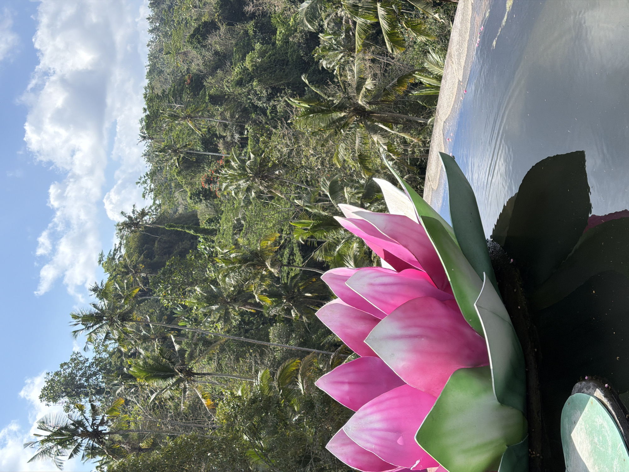 A large, artificial pink and green lotus flower floats on the edge of a pool of dark water. The flower has vibrant pink petals with white tips and green sepals. The water reflects the sky and the flower, and a few small pink flower petals are scattered on the surface and on the concrete edge of the pool. In the background, a lush, dense jungle with many palm trees stretches up a hillside under a bright blue sky with fluffy white clouds. This scene is located at a resort in Tampaksiring, Indonesia.