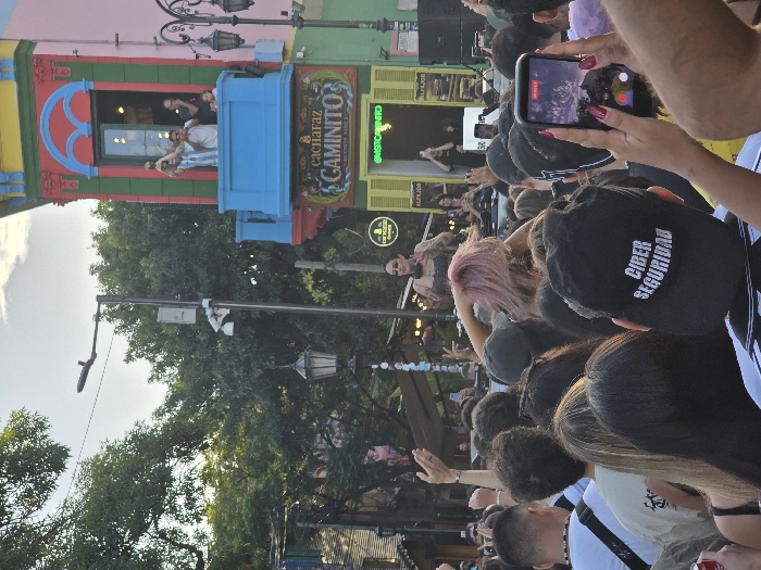 A crowd gathers in front of a building with colorful facades and street performers. In the background, a stage is set up for a performer wearing a dress. A woman in the foreground is holding up a cell phone to record the performance. The crowd appears tightly packed with people of different heights and hair colors. A hat in the foreground has the phrase "CIBER SEGURIDAD" printed on it. The sun illuminates the crowd, and trees frame the backdrop.