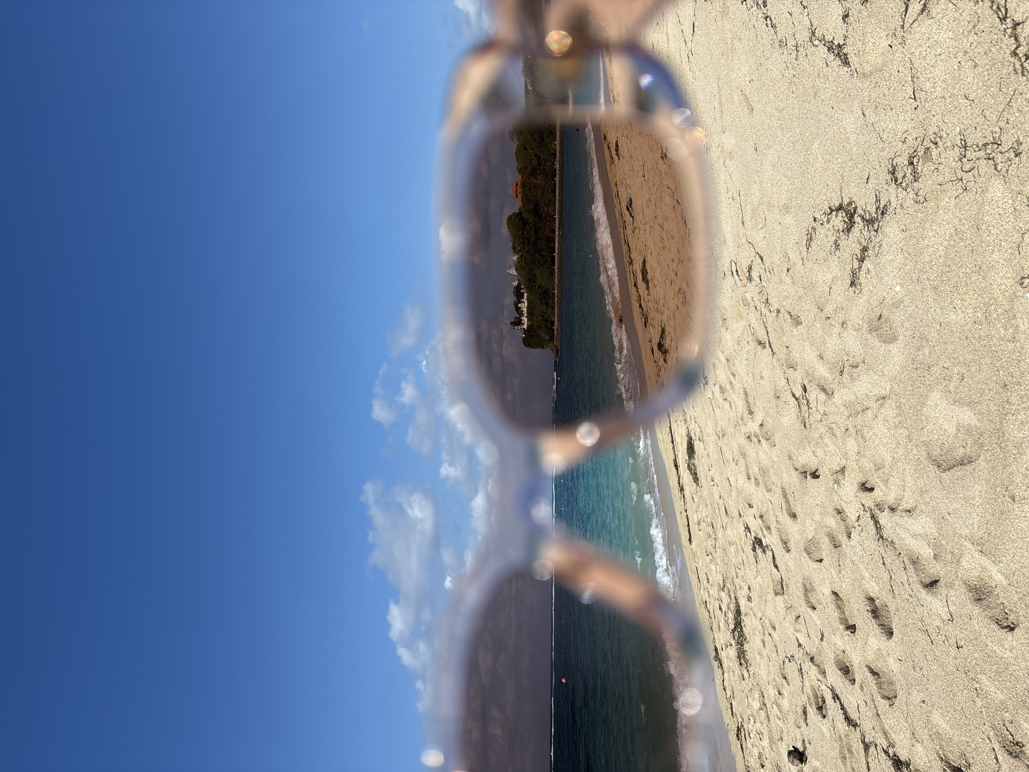 The image displays a vibrant beach scene viewed through a pair of light-framed sunglasses, rotated 90 degrees counter-clockwise.
When oriented correctly, the upper portion reveals a clear, bright blue sky with a few scattered white clouds. The lower portion features a sandy beach, marked with numerous footprints and some scattered dark seaweed. The ocean's edge runs diagonally through the middle, with gentle waves breaking on the shore.
The primary subject is the scene observed through the sunglasses' lenses. Through the lenses, a picturesque coastline is visible, featuring the turquoise ocean merging with a sandy beach. In the distance, across the water, a verdant landmass or resort area can be discerned, dotted with buildings, including one with a noticeable reddish-orange roof. The water appears calm and inviting. The sky seen through the lenses also shows some cloud formations.
Outside the lenses, the sky on the left is a deep blue, and the beach on the right is a golden sand color. The sunglasses frames are light-colored, and bright, circular reflections from the sun are visible on the lenses and edges, indicating strong sunlight. The overall atmosphere is one of a bright, sunny day. The scene evokes a tranquil holiday setting, consistent with a location such as Kuta Selatan, Indonesia. There is no visible text in the image.
