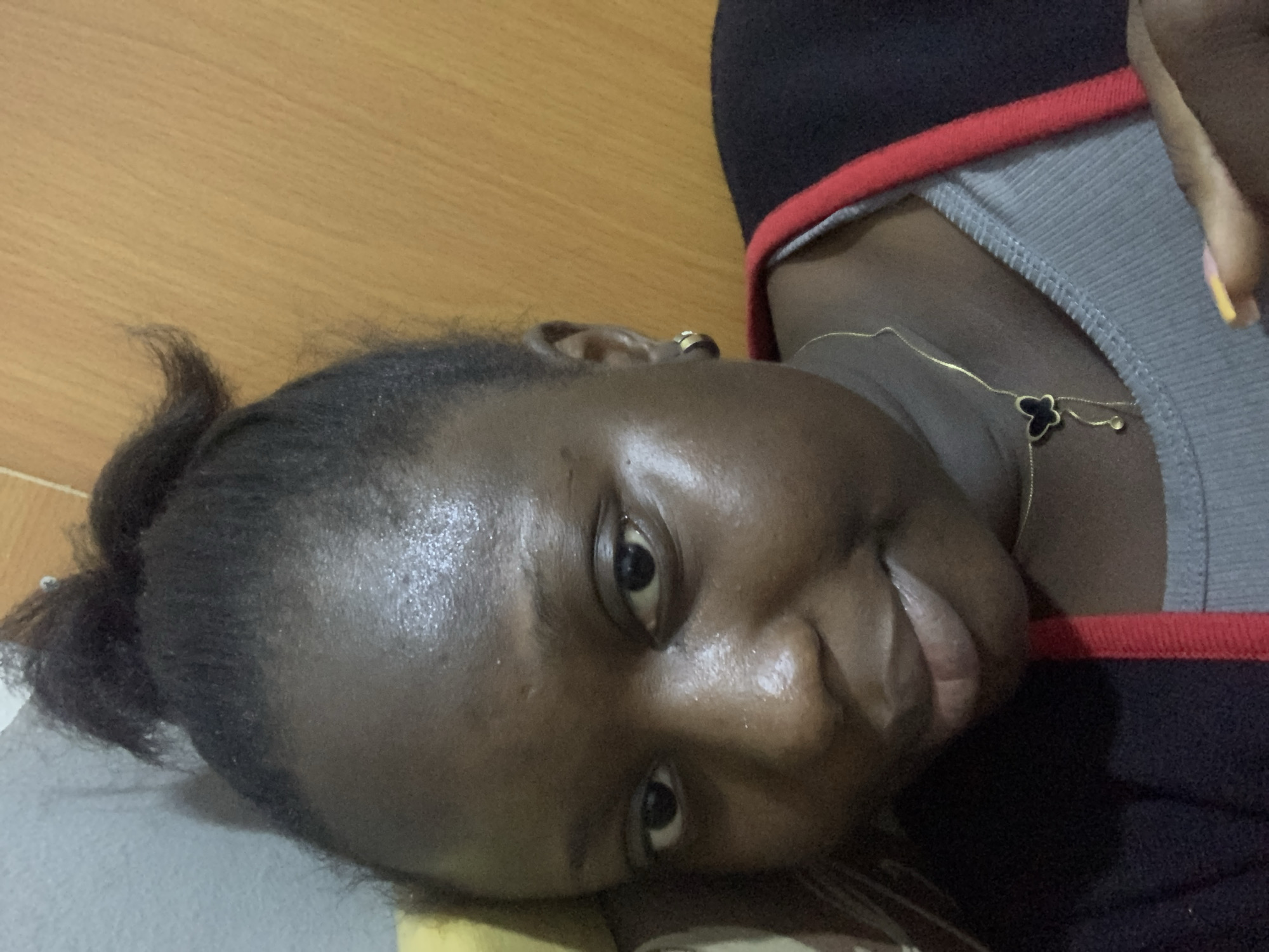 A young Black woman with her hair pulled up in a messy bun looks directly at the camera in a selfie. She is wearing a grey ribbed tank top under a black jacket with a red trim. A thin gold necklace with a black butterfly pendant is visible around her neck. She has a gold hoop earring in her left ear. Her skin appears slightly oily. The background consists of a grey wall to her left and a wooden surface to her right. The lighting suggests an indoor setting.