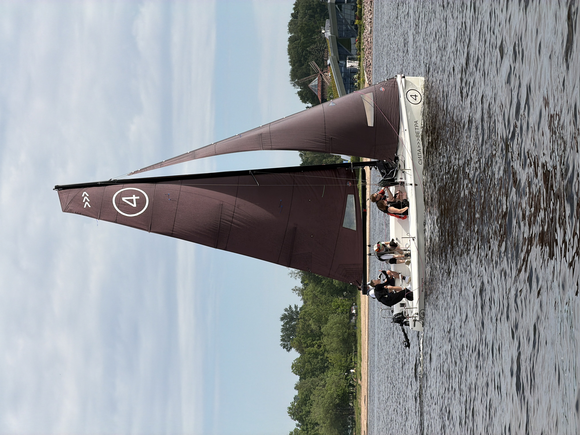 A sailboat with a large dark brown sail is sailing on a body of water. The sail has a white circle with a white number "4" inside, and two sets of three white chevrons above it. The boat is white and has several people on deck. On the side of the boat, in white lettering, is "СИЛА>>>ВЕТРА" and another white circle with a white number "4". The boat is moving away from the viewer, slightly to the right. In the background, there is a sandy beach, a line of green trees, a modern building with a glass facade and a green roof, and a windmill. The sky is overcast with some patches of blue. The water is choppy, with visible ripples.