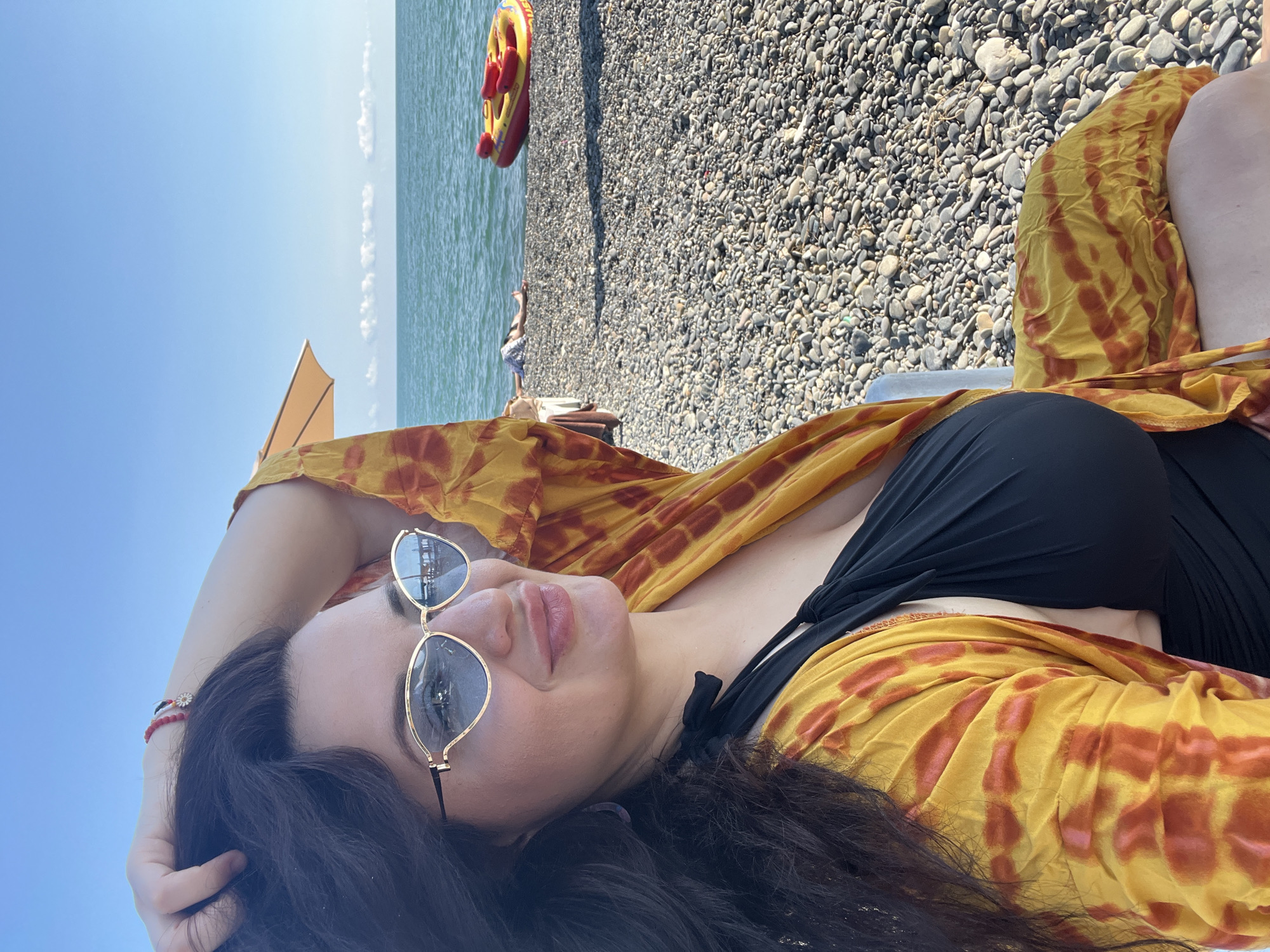 A woman with dark hair and sunglasses is sitting on a pebble beach in Sokhumi, Abkhazia. She is wearing a black swimsuit and a yellow and orange tie-dye cover-up. She is looking to the side and has her hand in her hair. The sea is in the background, and there is a yellow inflatable raft on the beach. The sky is blue with some scattered clouds.