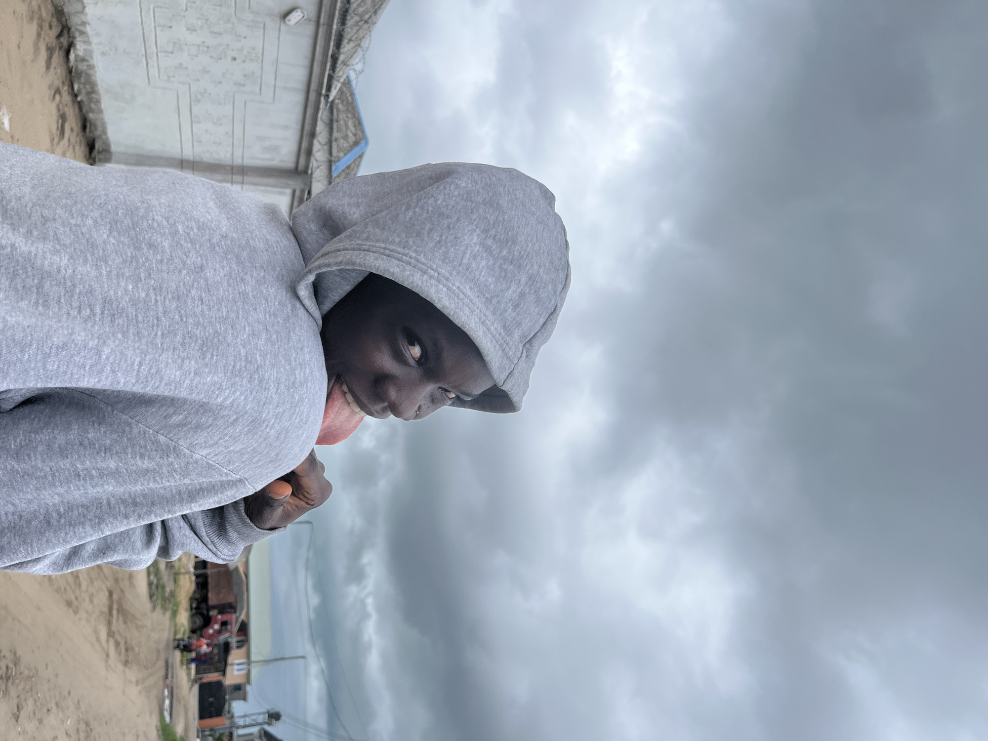 A young person wearing a gray hoodie with the hood up is shown in the photograph. The person's tongue is sticking out. The photo was taken at ground level on a dirt road outside the person. In the background are a few buildings with a sand-colored dirt road in front. Overhead, there are gray, heavily overcast skies. The image was taken in Badagry, Nigeria.
