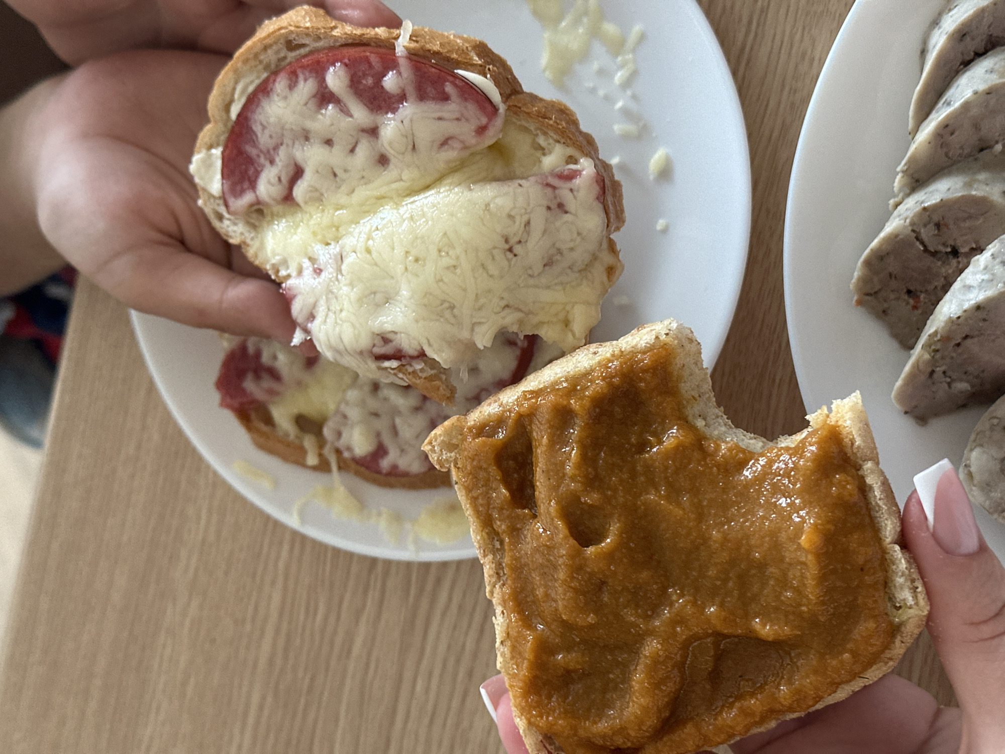 A person's hands are shown holding food items over a wooden table. In the foreground, a piece of bread with a thick, orange spread, possibly a vegetable puree, is held in a person's left hand. The bread has a bite taken out of it. In the background, on a white plate, are slices of bread topped with tomato and melted cheese. Another person's hands are holding one of these slices. A white plate with sliced sausage is visible in the lower right corner. The setting appears to be indoors, likely a home or cafe, with a glimpse of a blue fabric item in the upper part of the image. The overall scene suggests a casual meal or snack. The image does not contain any identifiable landmarks or clues that would suggest a specific location within Tallinn, Estonia.