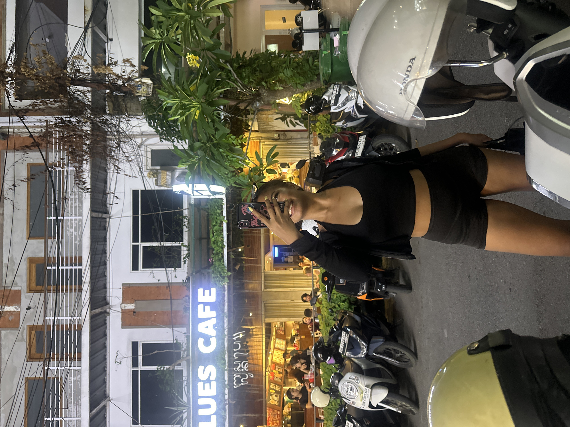 The image depicts a street scene in Denpasar, Indonesia, at night, in front of a cafe labeled "BLUES CAFE." A young woman, holding a smartphone up to take a selfie, stands in the foreground. She is wearing a black crop top and black shorts. Behind her, several parked motorcycles are visible. In the background, the "BLUES CAFE" sign is illuminated, and patrons are seated inside the establishment. The building behind the cafe has multiple stories with balconies and windows. Trees and foliage are present, and electrical wires are strung across the scene, typical of urban environments.