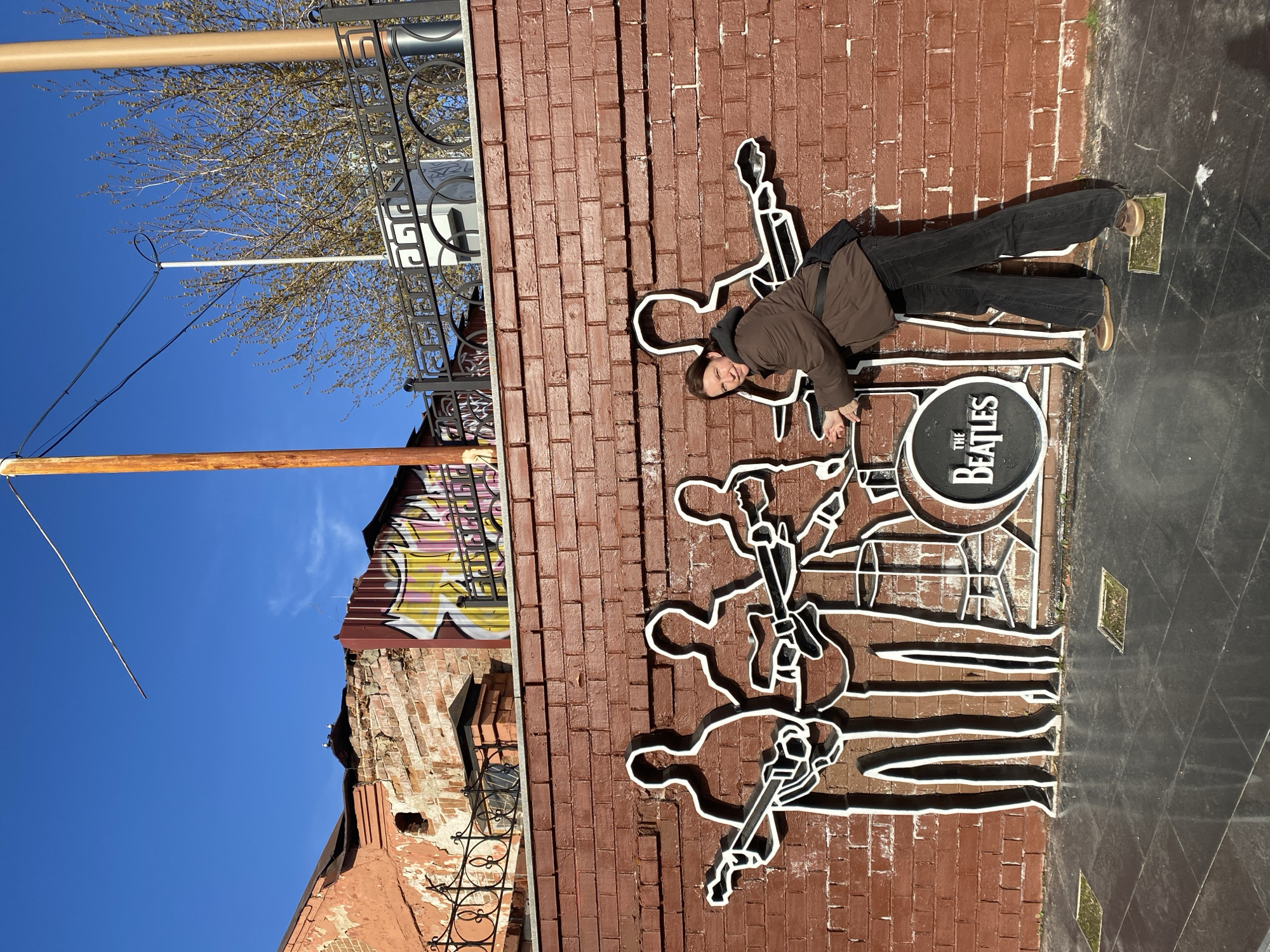 The image shows a person in front of a brick wall with a depiction of the Beatles. The person is leaning against the brick wall, with the Beatles artwork seemingly attached to it. The artwork features white outlines of the band members and a drum set with "THE BEATLES" written on it. The brick wall appears to be part of a building facade. To the left of the wall, there is a glimpse of another building with some graffiti and a wrought-iron fence. In the background, a blue sky is visible with some trees. The scene is located in Yekaterinburg, Russia, as indicated by the Beatles monument on display.