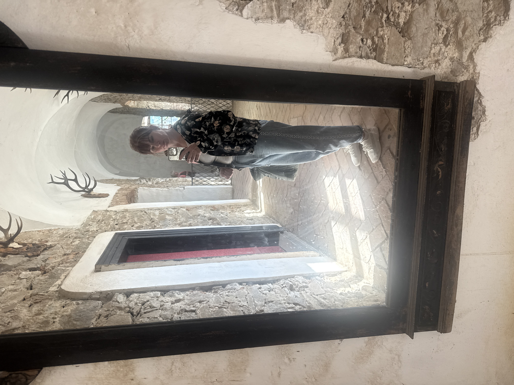 The image shows a person reflected in a large, ornate mirror mounted on a stone wall in what appears to be the interior of a historic building. The person is holding a phone and taking a picture of themselves. The reflection reveals a long corridor with a vaulted ceiling, architectural details like antlers mounted on the wall, and a small window. The context clues provided lead to a conclusion that this image was taken in or near the city of Chynadiiovo, Ukraine, likely inside a castle or other historical site.