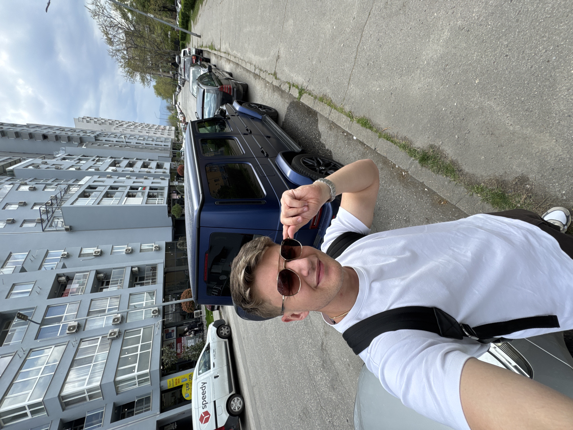 The image, taken as a selfie and rotated 90 degrees clockwise, captures a young man on a street in what appears to be Plovdiv, Bulgaria. The man, positioned in the foreground, wears a white t-shirt and dark sunglasses, which he is adjusting with his right hand. He has light-colored hair and a faint smile, suggesting a relaxed mood. A black strap, possibly from a bag, crosses his chest.
Behind him, a dark blue Mercedes-Benz G-Wagen is prominently parked on the side of an asphalt road. Other cars are lined up along the street, including a dark sedan and a white delivery van with the "Speedy" courier service logo visible on its side. The sidewalk is made of concrete with some patches of grass growing along the curb.
In the background, tall, multi-story buildings with numerous windows and outdoor air conditioning units line the street, characteristic of urban residential or commercial architecture. On one building, Cyrillic text is visible on signs; one sign appears to read "СВЕТЪТ ВОДНИ СТЪПКИ" (The World of Water Steps/Glass) and another has "ЗАР" and phone numbers. The sky is partly cloudy, indicating daytime, likely morning or afternoon. Sparse trees with budding leaves are visible further down the street, suggesting spring or early summer weather.