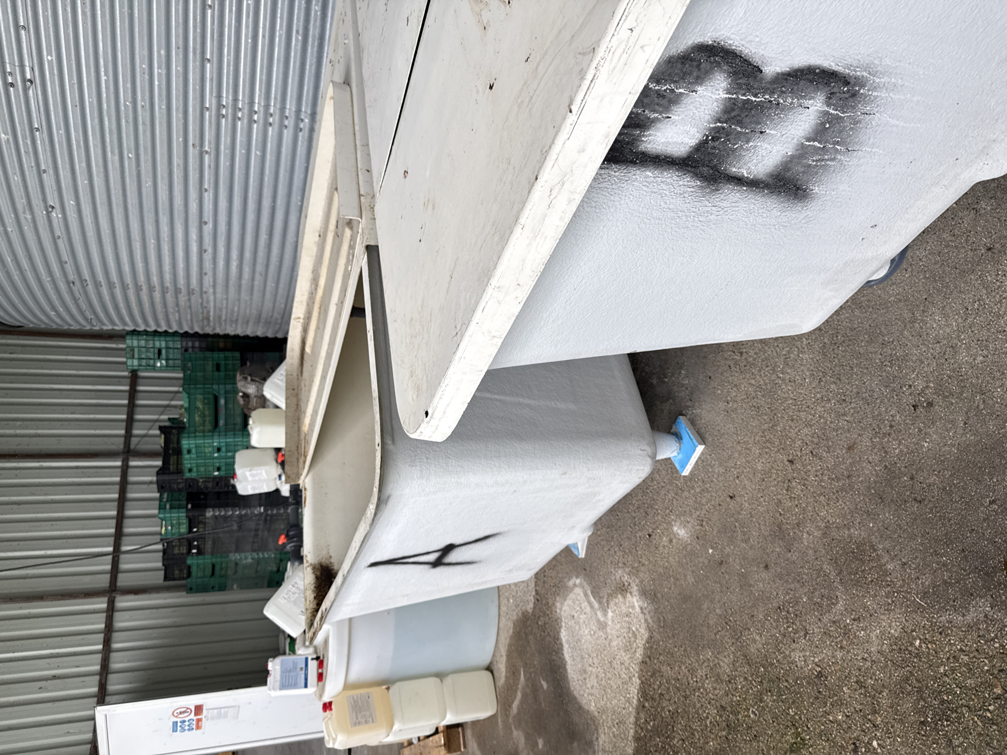The image depicts an industrial or storage area, likely inside or adjacent to a building with a prominent corrugated metal wall. No people are visible.
The scene is dominated by several large, light-grey or off-white industrial containers, possibly tanks or tubs, made of plastic or fiberglass. One such container is featured centrally with its hinged lid open, revealing an empty interior. On its side, the number "200" is clearly spray-painted in black. Another similar container is positioned below it, marked with a stylized black "4." A blue plastic cap or plug is visible on the side of one of the containers near the bottom.
In the background, a large quantity of green and black plastic crates are stacked against the corrugated metal wall. Various smaller white and yellowish plastic containers and bottles are placed on the ground next to the larger tanks. A tall, white appliance resembling a refrigerator or cabinet, featuring some small, mostly illegible labels (e.g., "Jura," "900," "000"), is partially visible on the far left.
The ground is a grey concrete or asphalt surface, appearing slightly damp in places. The lighting suggests it is daytime, possibly under an overcast sky, creating soft, even illumination. The overall setting indicates a commercial or industrial facility in Stara Zagora, Bulgaria, used for storage or handling of materials. No specific activity or event is taking place; the scene portrays an organized but utilitarian storage environment.