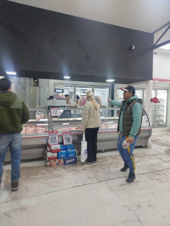 The image captures an interior view of a butcher shop or meat department within a supermarket located in Vertientes de Pedemonte, Argentina. In the center, a long, refrigerated glass display case is filled with various raw meat products, including cuts of poultry and red meat. Prominently displayed on the counter are price signs, such as one marked "$455" for "PATA DE M." and two on the floor for retail beverage displays, "$8935" for Andes beer and "$7999" for Quilmes beer. A digital scale is visible behind the counter.
Three customers and one employee are present. Behind the counter, a male employee, wearing a dark cap and an apron, gestures with his right hand, facing the customers. In front of the counter, a female customer with blonde hair, seen from behind, wears a light-colored jacket and dark pants, holding a white shopping bag. To her right, a male customer, wearing a black baseball cap, a green vest over a light-colored long-sleeve shirt, and blue track pants with yellow stripes, holds a clear plastic bag containing meat in his left hand and points with his right hand. To the far left, another male customer, seen from behind, wears a green fleece jacket and blue jeans.
The environment includes a light-colored concrete floor, a dark overhead bulkhead, and bright overhead lighting. In the background, additional self-service refrigerated units are visible, along with signage partially reading "AUTOSERVICIO" and "CONSULTAS Y PEDIDOS 2616RP." A single security camera is mounted on the dark bulkhead above the counter.
