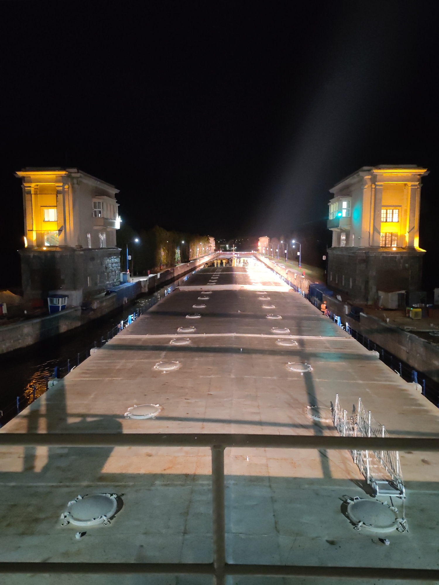 Captured at night, the image presents a straight elevated concrete structure flanked by waterway locks in Водник Снт, Russia. Two illuminated, multi-story control towers bookend the structure on either side. The concrete surface features several circular access points and a metal ladder. Further down the structure, a group of people wearing bright attire can be seen clustered near lighting fixtures. The environment is dimly lit, with street lights and distant buildings visible.