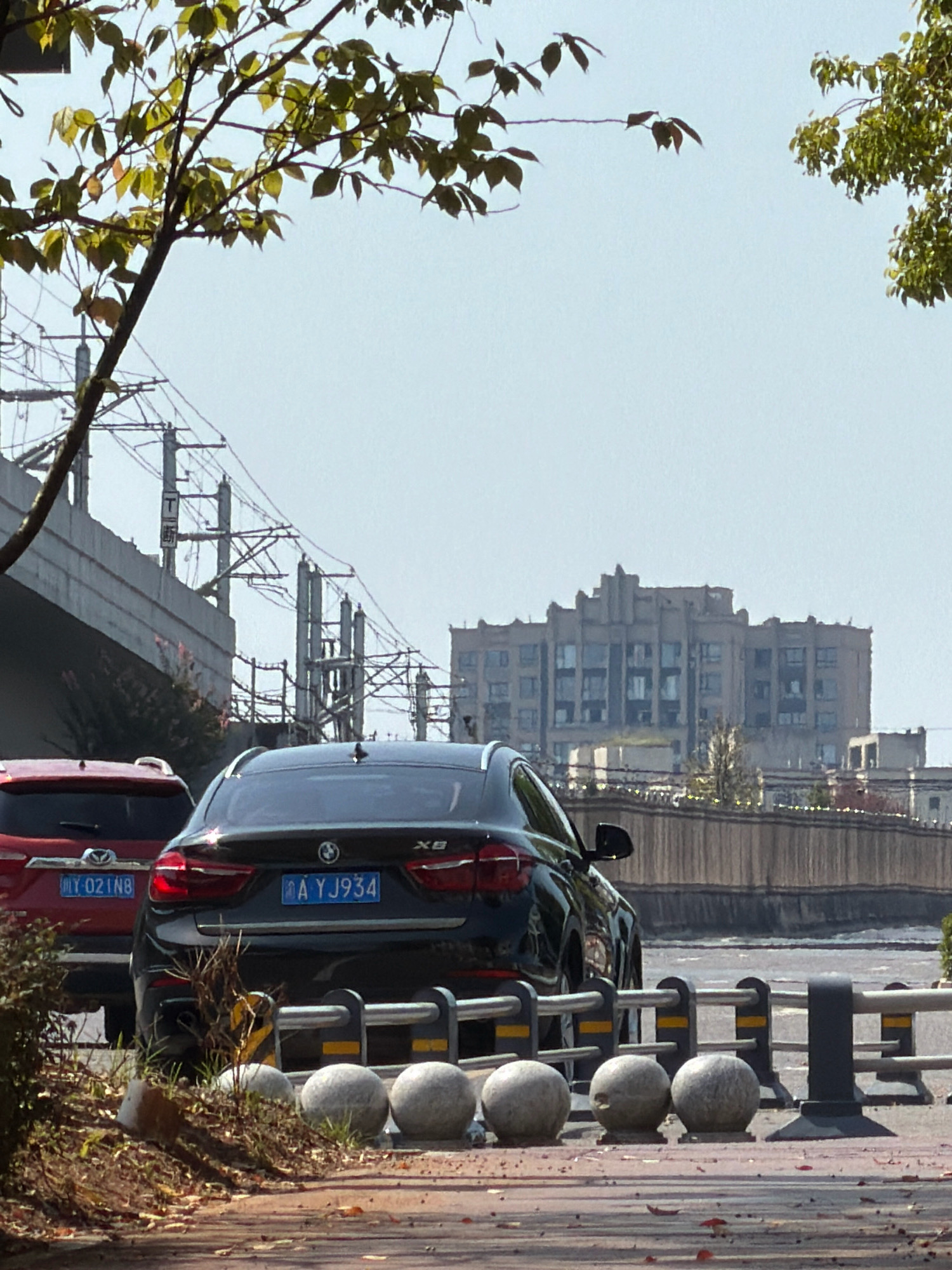 In Jiangjin, China, a black BMW SUV is parked on a paved surface. The car has a license plate that reads "A YJ934". To the left of the BMW is a red SUV with a license plate reading "Y 021N8". A tree with green leaves is in the foreground, partially obscuring a bridge and overhead train tracks. In the background, a large, multi-story building with a distinctive architectural style is visible. The sky is clear and pale blue. The scene appears to be a street or parking area, with a barrier in front of the parked vehicles.