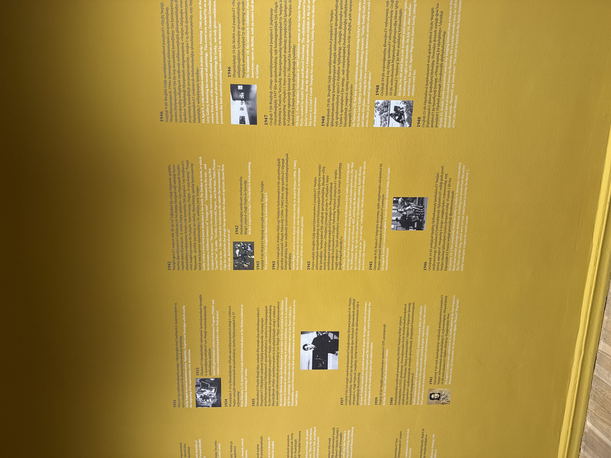The image shows a timeline on a wall, possibly in a museum or gallery in Yerevan, Armenia. The timeline features text (in both Armenian and English) and photographs related to the life and career of Arshile Gorky, an Armenian-American painter. The timeline spans from 1931 to 1948 and details Gorky's artistic development, exhibitions, personal life, and eventual suicide. The text provides historical and biographical context alongside black-and-white images of Gorky, his family, and his artwork.