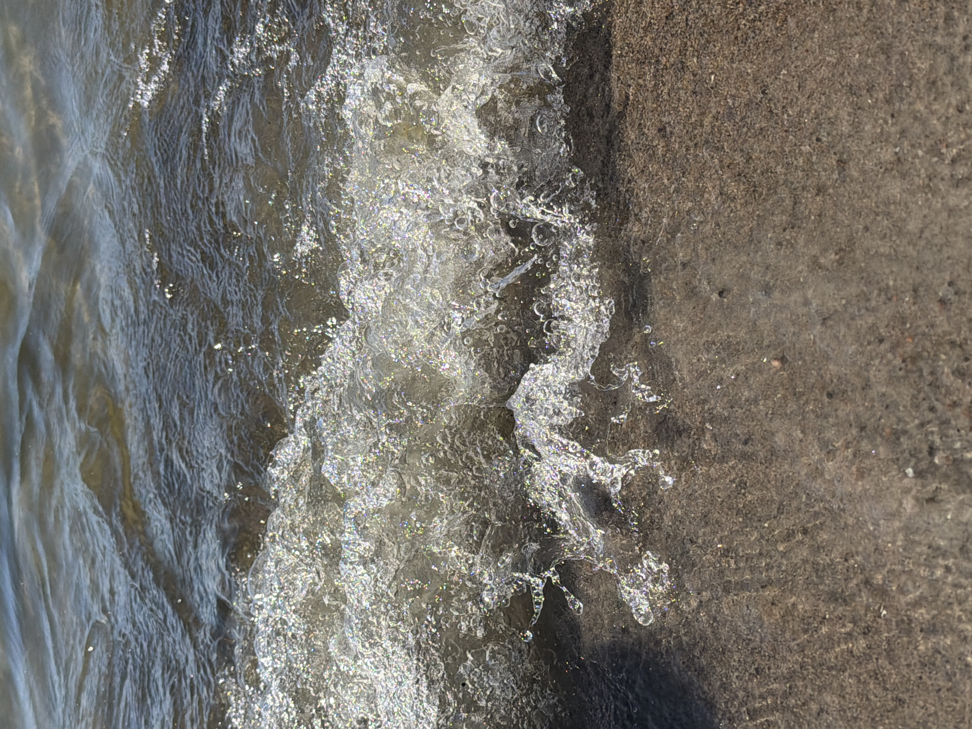 A close-up, eye-level shot shows a small wave gently lapping onto a sandy shore. The water is clear and reflects the sunlight, creating a sparkling effect on the crest of the wave. The sand is a light brown color, with small pebbles and shells visible. The background shows the murky depths of the water, with ripples and light reflecting off the surface. The scene appears to be a natural body of water, possibly a lake or the sea, with no human presence visible. The lighting suggests it is daytime. The specific location in Tallinn, Estonia cannot be determined from the image alone.