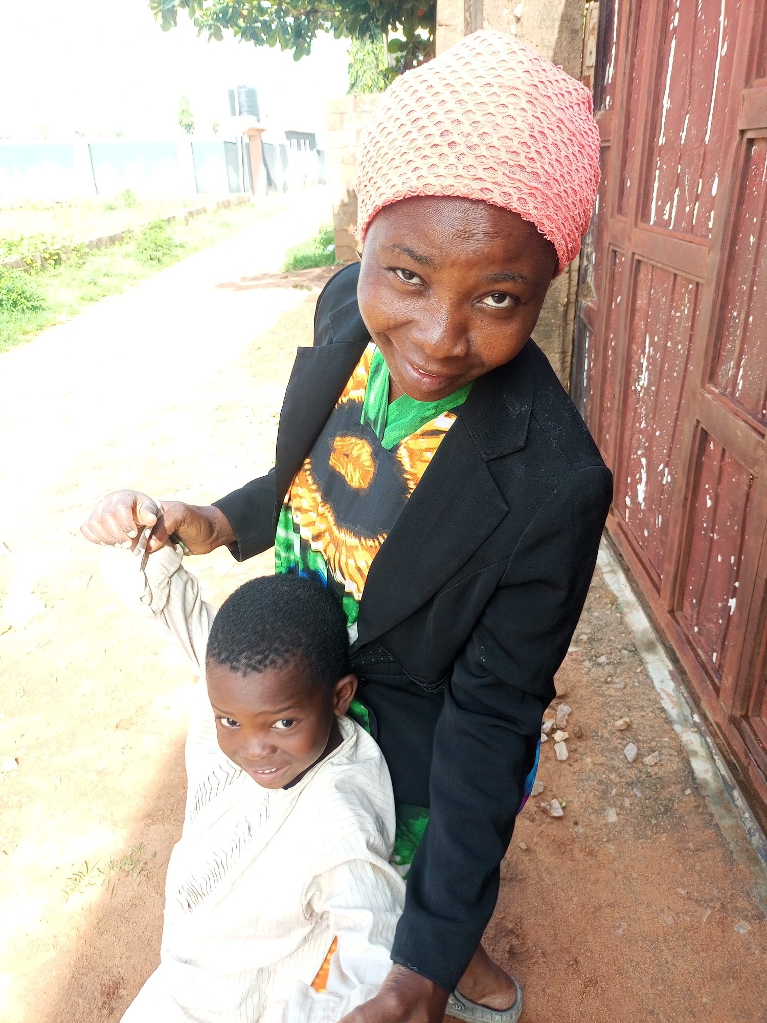 An eye-level, medium shot captures a Black woman and a young Black boy in Tempure, Nigeria. The woman, positioned to the right, wears a coral-pink headscarf and a black blazer over a green, orange, and black patterned shirt. She is holding the boy's left wrist with her right hand. The boy, slightly to the left and lower in the frame, faces the camera, dressed in a light-colored, striped garment. He is looking at the camera with a subtle smile. Behind them, a reddish-brown wooden door with vertical panels and white splotches is visible on the right. The ground is composed of red dirt and small stones. In the background, a dirt path leads away from the subjects, bordered by sparse green vegetation and a pale fence. Trees with green foliage are visible at the top of the frame. The lighting suggests it is daytime.
