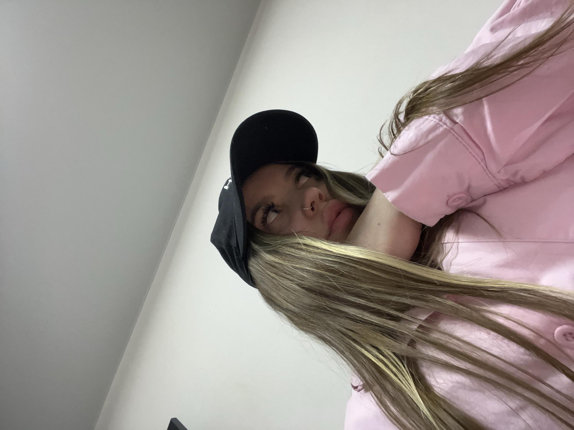 A low-angle, close-up portrait features a young person, likely female, indoors. The subject has long, straight hair, appearing light brown with distinct blonde highlights, and wears a black baseball cap with some unidentifiable white text or logo on the front. A pink collared shirt or jacket is visible, partially obscured by their hair. The person has a small hoop nose piercing. Their gaze is directed upwards and slightly to the right, with a pensive or slightly bored expression, characterized by a subtle pout and slightly parted lips. The background is composed of plain, light-colored walls, indicating an indoor environment. While the location context is Krasnodar, Russia, the image itself does not contain specific geographical markers.