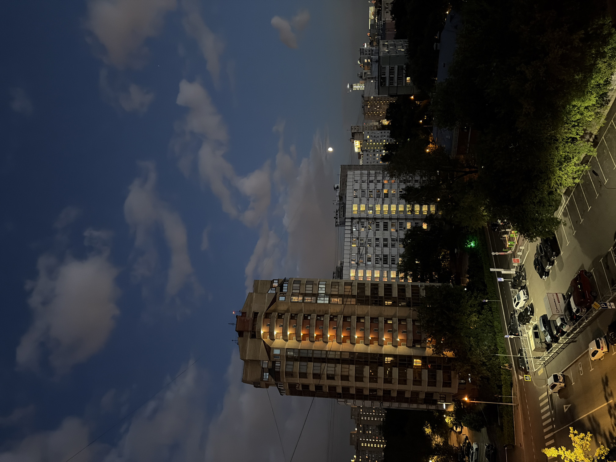The image depicts an urban night scene in Moscow, Russia, likely taken from a high vantage point overlooking a residential area. A tall, multi-story apartment building with illuminated windows dominates the left side of the frame. Its facade is a mix of concrete and glass, with balconies showing warm interior lights. To the right, several other apartment buildings of varying heights are visible, also featuring lit windows, suggesting the time is evening or night. The sky is a deep blue with scattered white clouds, and a crescent moon is visible in the upper right quadrant. In the foreground, a street and a parking lot are illuminated by artificial lights. Several cars are parked in the lot, and a pedestrian walkway with crosswalk markings is visible. Trees and greenery are interspersed throughout the scene, adding a natural element to the urban landscape. The overall impression is a serene, lived-in city at dusk or after dark.