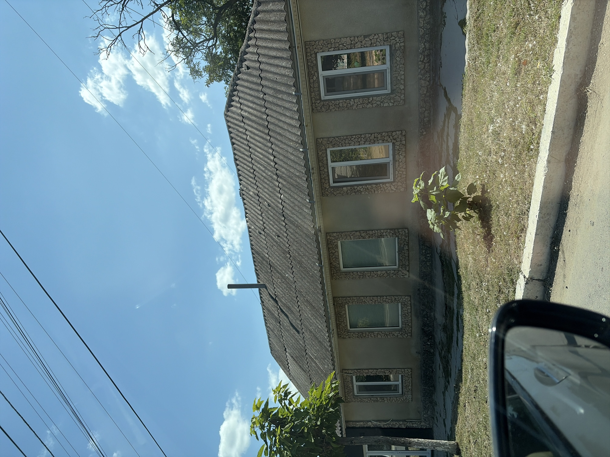 The image shows a single-story house with a dark, corrugated roof and a light-colored facade. The windows are framed with decorative stone. There is a chimney pipe on the roof. In front of the house, there is dry grass with some scattered white fluff, a small green plant, and a paved area. A tree with green leaves is visible to the left of the house. The sky is blue with some white clouds. Overhead power lines are visible in the upper part of the image. The reflection of a car's side mirror is in the lower left corner. This scene is located in the city of Criuleni, Moldova.