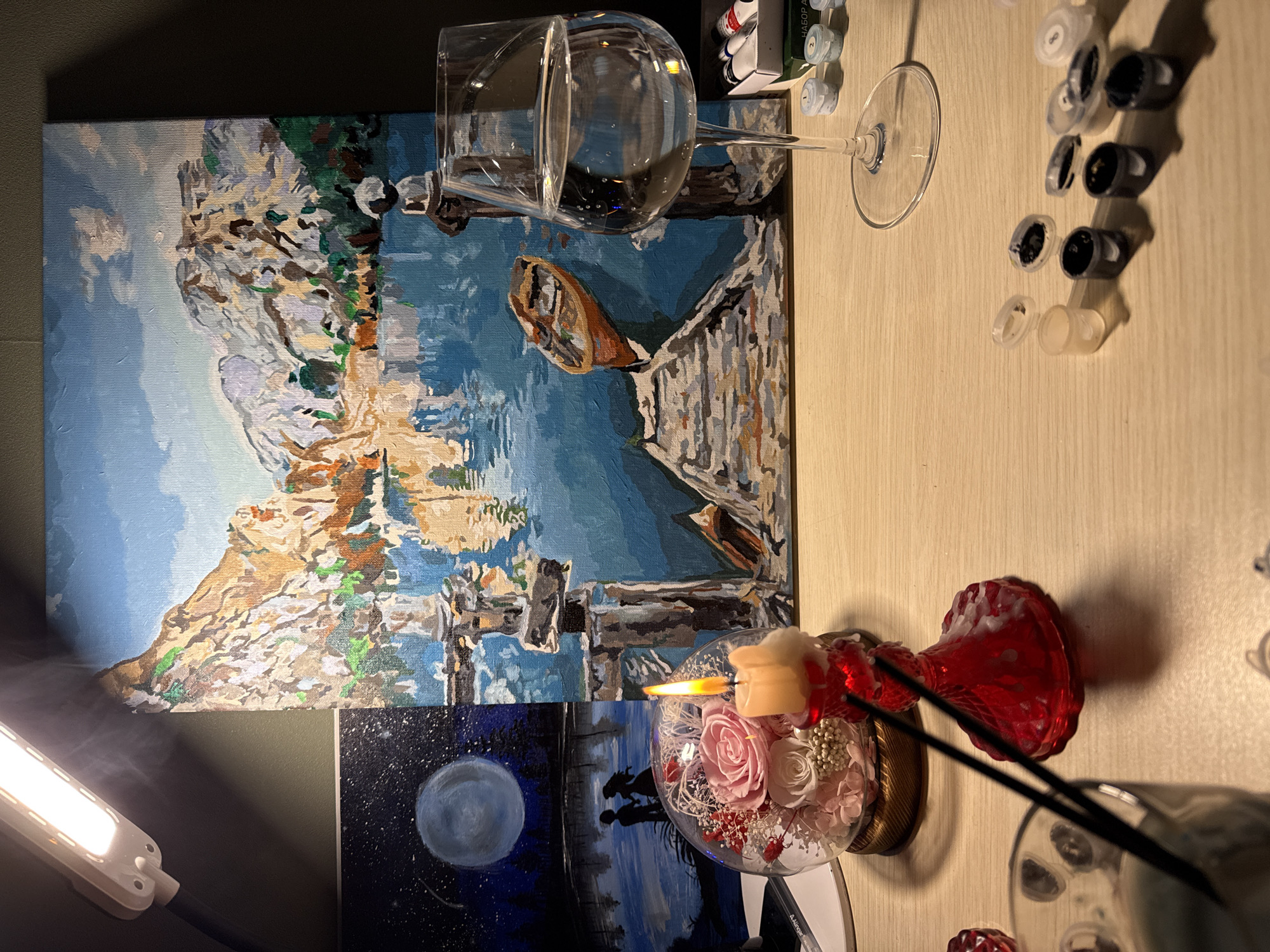 The image depicts a still life setup on a wooden surface, possibly a desk or table, with a focus on art supplies and decorative elements. In the background, two paintings are visible. The larger painting, centrally placed, is an abstract landscape featuring mountains, a body of water reflecting the sky, a small boat, and a wooden dock. The smaller painting to the left shows a night scene with a large moon, stars, a lake, and silhouettes of two figures holding hands.
In the foreground, a lit candle in a red, ornate candle holder casts a warm glow. To the left of the candle is a glass dome containing preserved flowers, primarily pink and white roses, with some red accents. Next to the dome, two black chopsticks are partially submerged in what appears to be a container of liquid.
To the right of the main painting, a tall wine glass filled with clear liquid, likely water, stands prominently. Bubbles are visible within the liquid. Beside the wine glass, a box of small paint pots and tubes is arranged, suggesting the paintings are part of a paint-by-numbers kit. Some of the paint pots are open, revealing different colors.
An overhead lamp is positioned above the paintings, illuminating the scene. The overall ambiance suggests a creative or artistic endeavor, perhaps a moment of relaxation or work. The location in Cheboksary, Russia, is not discernible from the visual elements in the image.