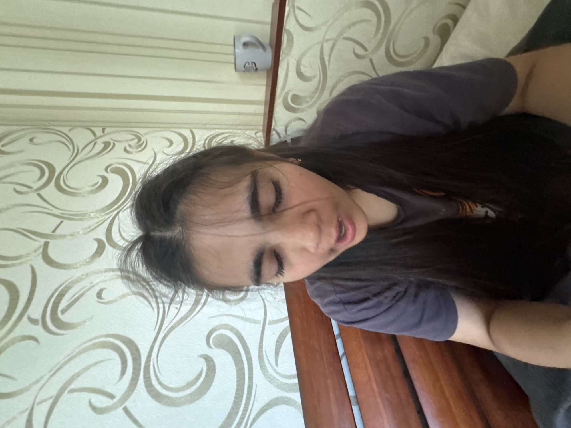 A young girl with dark hair and closed eyes is looking down with her mouth slightly open. She is wearing a dark t-shirt. The background consists of wallpaper with a swirling pattern and vertical stripes. To the right, a shelf holds a white mug. The image appears to have been taken indoors, possibly in a bedroom, in Poltava, Ukraine.