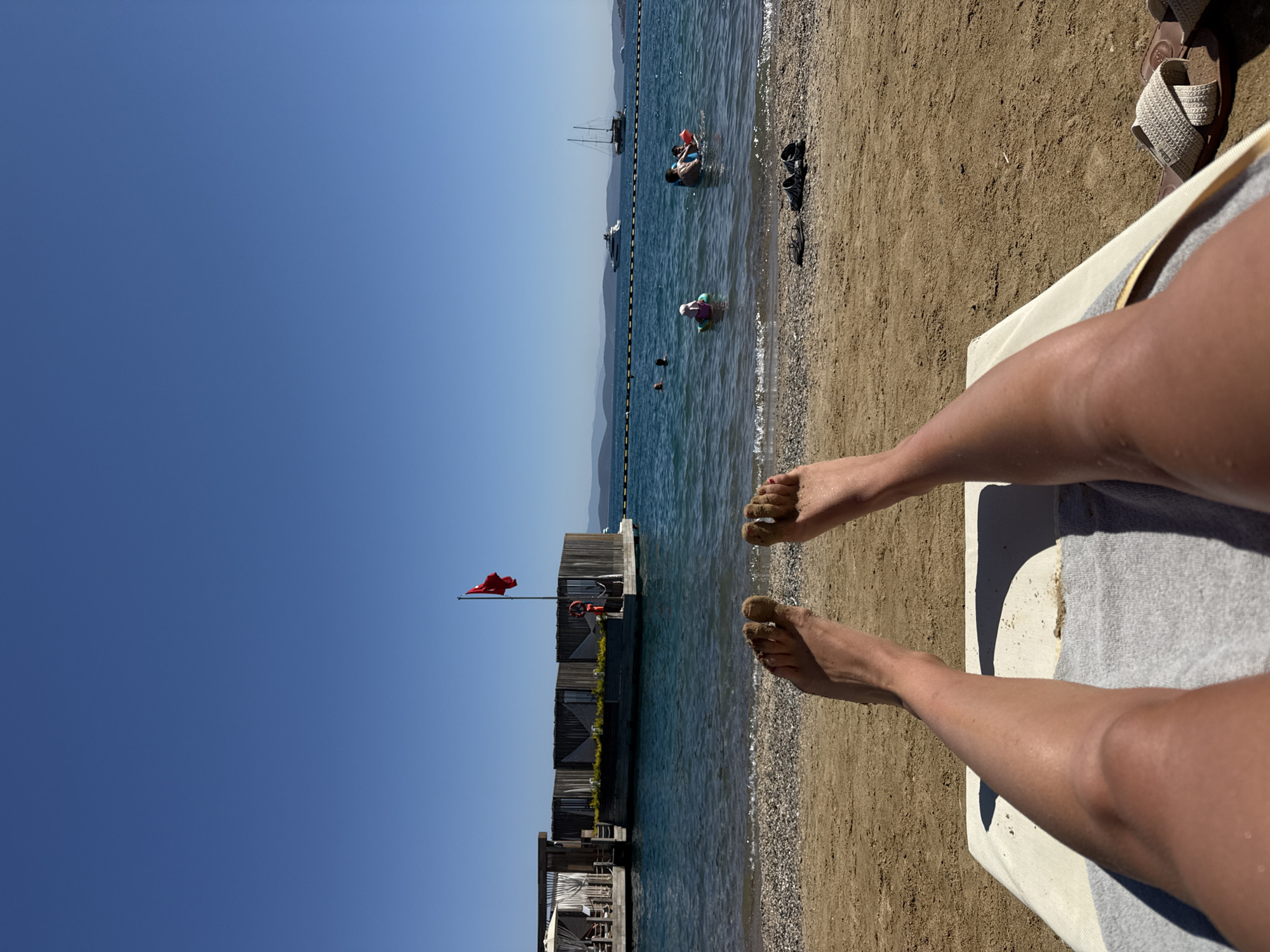 The image depicts a sunny day at a beach in Bodrum, Turkey. In the foreground, a person's legs and feet, dusted with sand, are visible as they recline on a lounge chair. The beach is sandy with some pebbles near the water's edge. The sea is calm and blue, with a few buoys marking a swimming area. In the water, several people are enjoying themselves, including children in inflatable rings and adults swimming. A floating wooden structure with several cabanas and a Turkish flag is situated in the sea, to the left of the frame. In the distance, boats, including a yacht and a sailboat, are visible on the horizon, with hills forming a backdrop. The sky is clear and a deep blue.