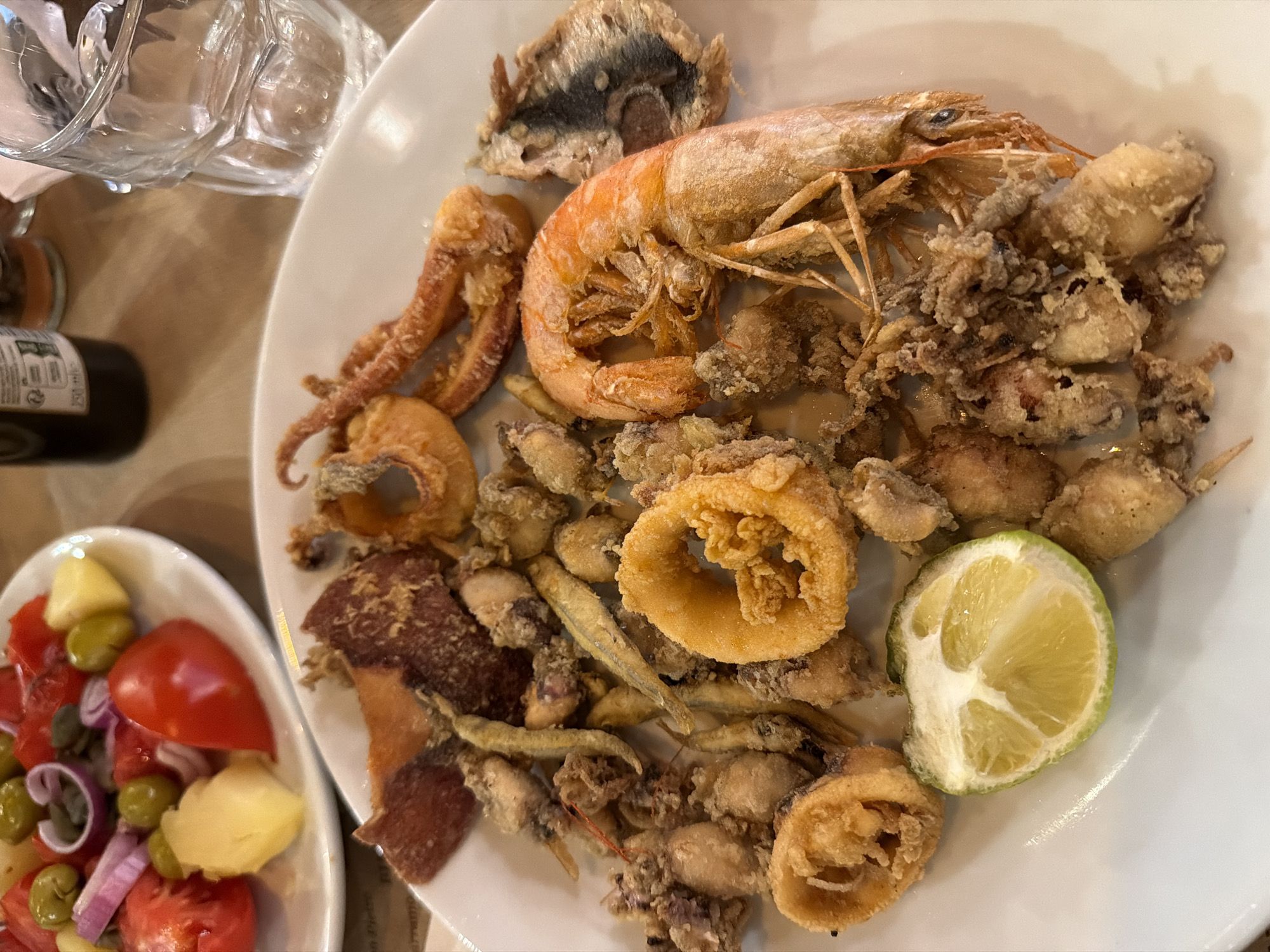 A plate of fried seafood is presented on a white plate, accompanied by a slice of lime. The seafood includes calamari rings, small fish, shrimp, and what appears to be octopus. In the background, another plate holds a tomato and olive salad. A glass of water and a bottle of dark liquid, possibly wine or olive oil, are visible on a wooden table. The setting is a restaurant in Palermo, Italy, suggested by the cuisine and the overall ambiance. The lighting is warm and natural, highlighting the textures and colors of the food.