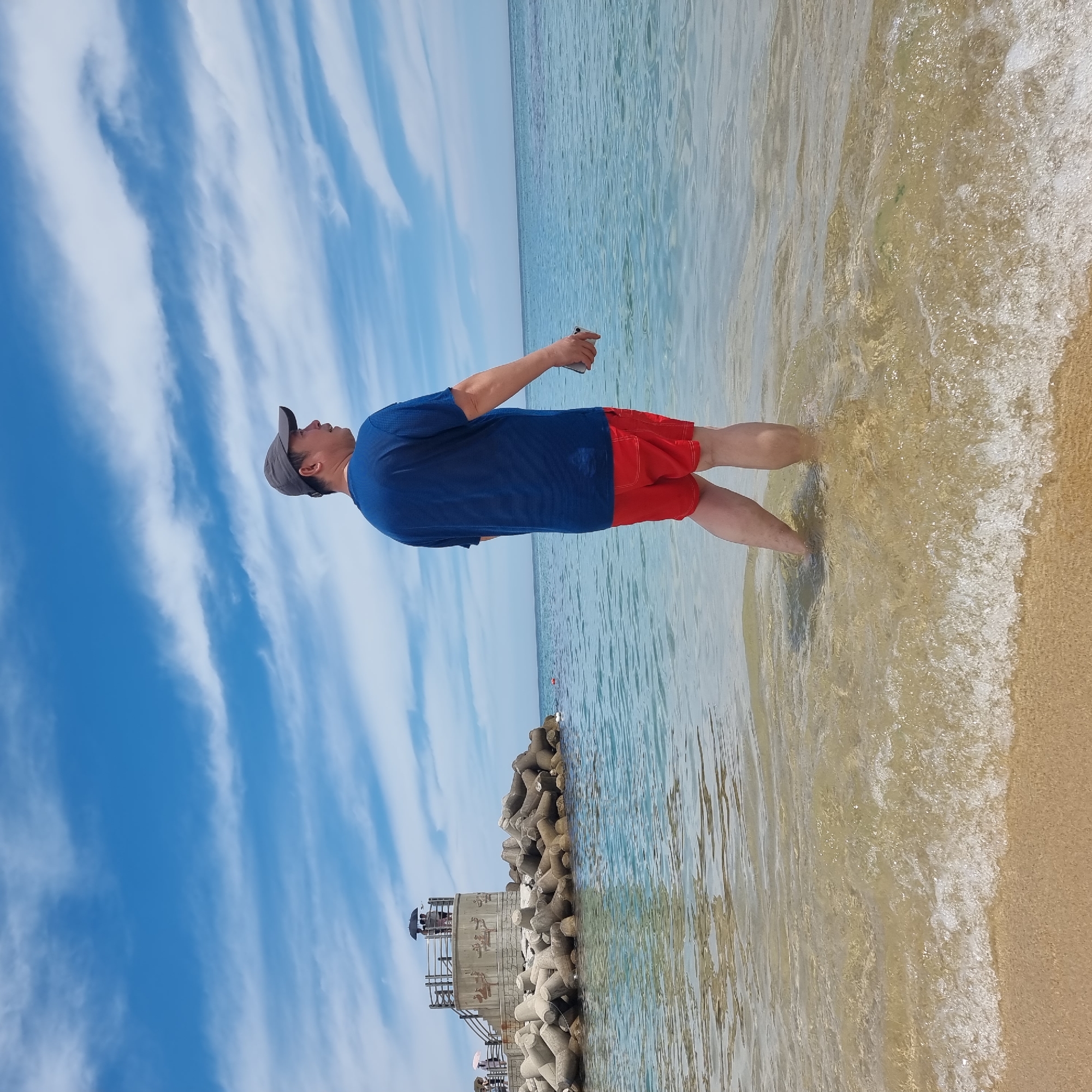 A man wearing a blue t-shirt and red shorts stands in shallow water at a beach. He is facing away from the camera, looking out at the ocean under a blue sky with white clouds. A grey baseball cap is on his head. He is holding a mobile phone in his right hand. In the background, there is a breakwater made of concrete tetrapods and a small observation deck with a few people on it. The sea is clear and calm, with gentle waves lapping at the shore. This scene is likely located at a beach in Gangneung-si, South Korea.
