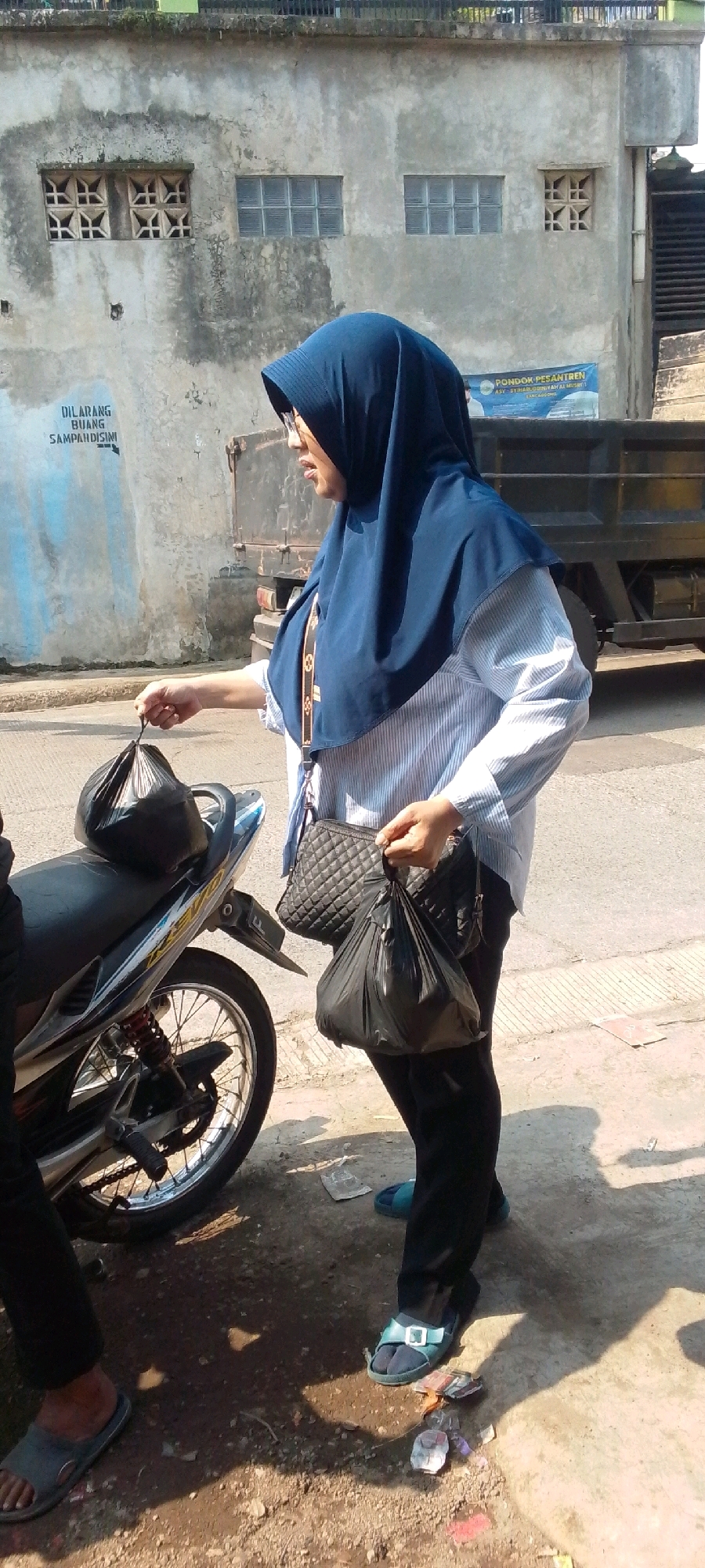 An Indonesian woman wearing a dark blue hijab, glasses, and a light blue and white striped long-sleeved shirt is standing outdoors. She is holding two black plastic bags, one in her right hand and another tucked under her arm. A quilted black handbag is slung over her left shoulder. She is wearing black pants and blue sandals. To her left, a motorcycle is parked, with one of the black plastic bags resting on its seat. The ground is a mix of pavement and dirt, with some scattered debris. In the background, a weathered concrete building with windows is visible, along with a large truck. A sign on the building reads "DILARANG BUANG SAMPAHISINI," which translates to "DO NOT LITTER HERE." Another sign above the truck reads "PONDOK PESANTREN ASY-SYUHAIBIYAH AL-MUSLIHI LANCAGGONG." The scene appears to be a street in Cilaku, Indonesia, on a sunny day.