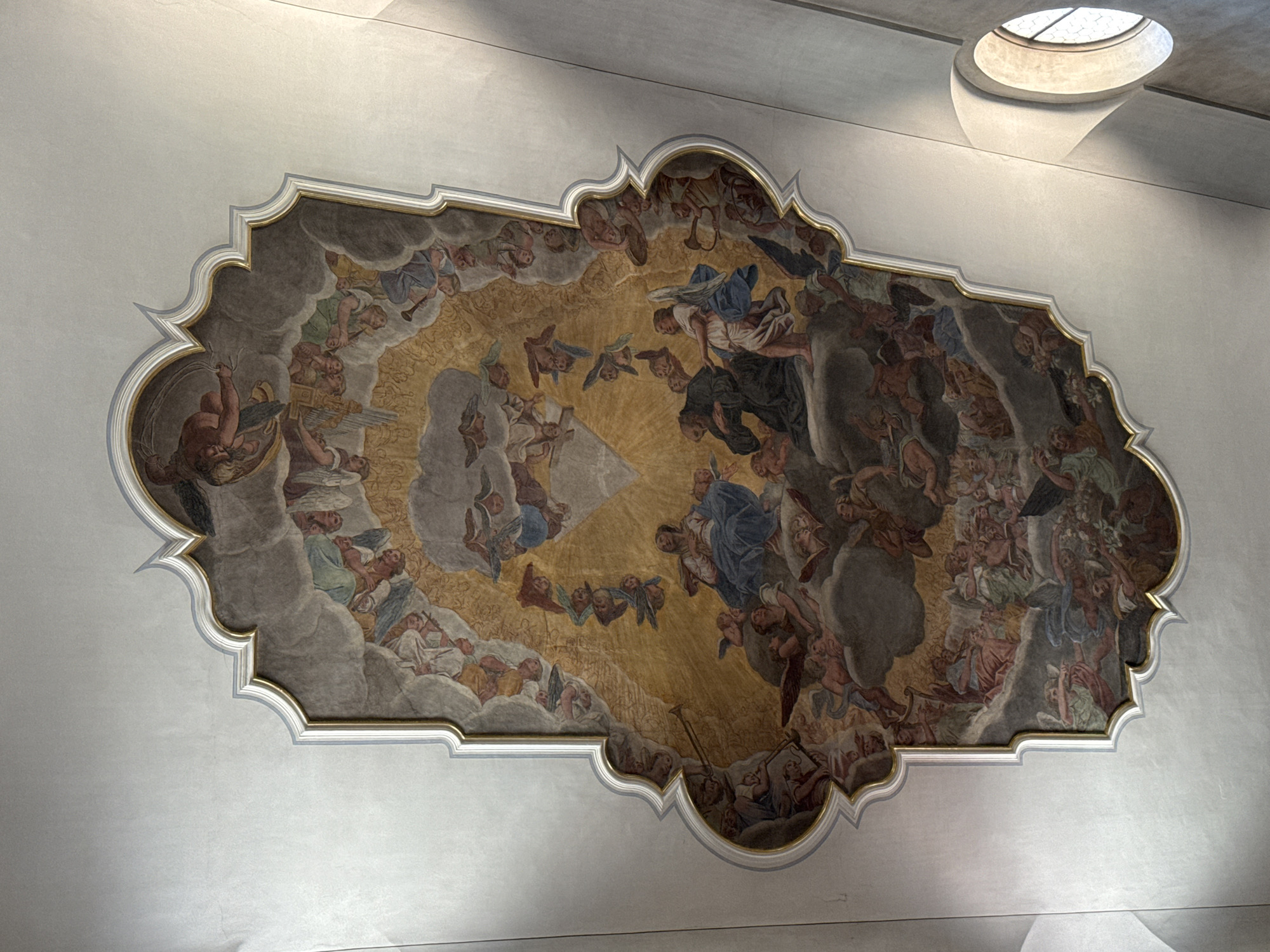 This is a photograph of a fresco painted on a ceiling, likely in the Jesuit Church in Lucerne, Switzerland. The fresco depicts a religious scene with numerous figures, primarily angels and humanoids, gathered in a celestial setting.
In the center of the composition, a woman in blue is seated and looking upwards, surrounded by other figures. To her left, a robed man with a beard is kneeling, with another winged figure in white reaching towards him. Scattered throughout the fresco are many winged figures, some appearing to be cherubs or putti, as well as other human figures in classical attire. Some figures are playing musical instruments, such as a trumpet and a lyre. The overall arrangement suggests a divine gathering or ascension.
The fresco is framed by an ornate, scalloped border with gold accents. The ceiling itself is white and appears to be slightly curved. To the right of the fresco, a rounded window with a latticed pane is visible, emitting light that casts shadows on the ceiling. The lighting in the scene suggests an interior space illuminated by natural light.